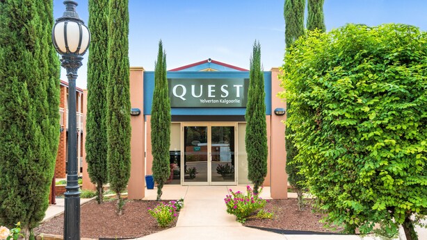 Exterior de un hotel llamado 'Quest', con una entrada despejada, un camino que lleva a la puerta y jardines bien cuidados, que incluyen árboles podados y macizos de flores.