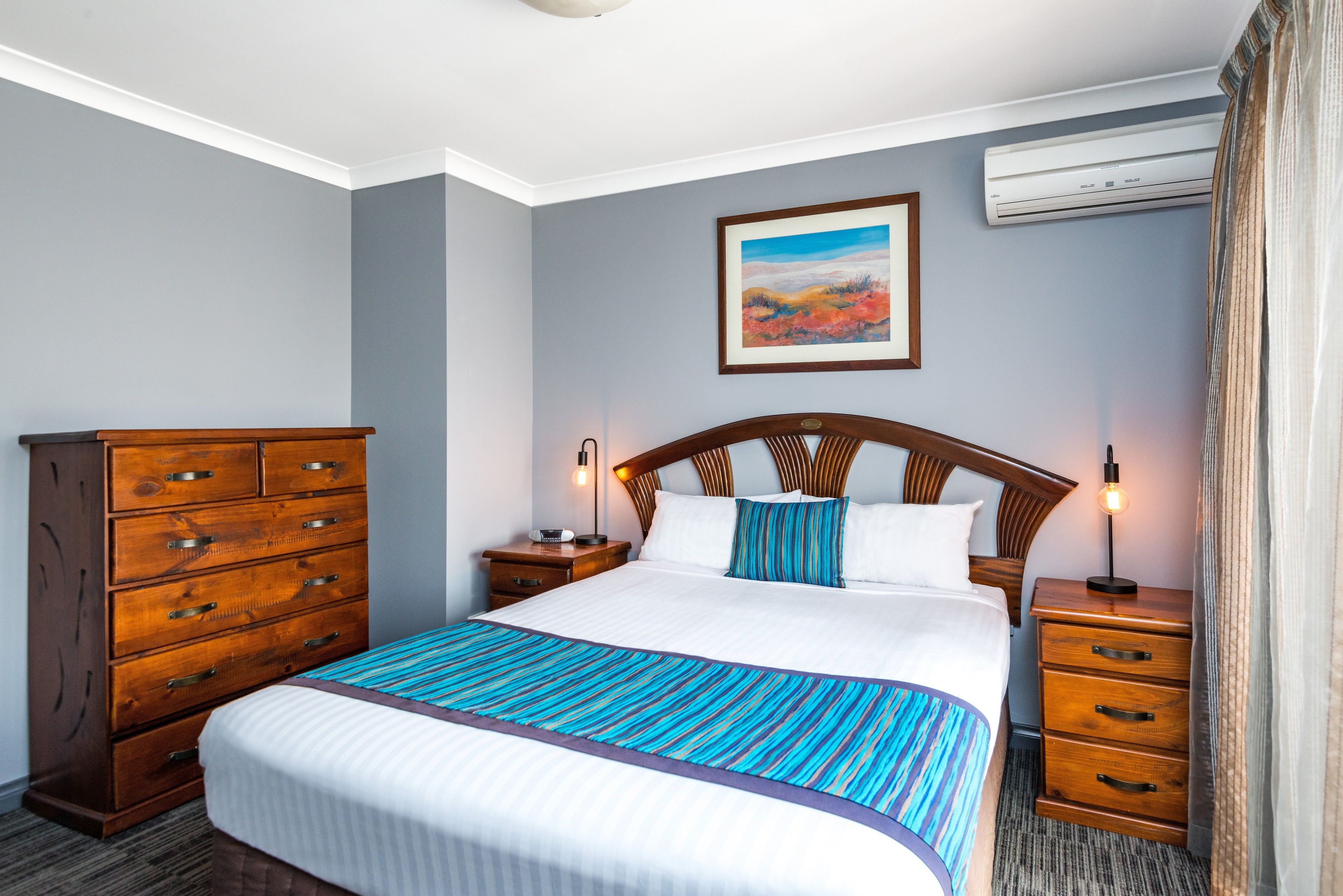 A hotel room with a large bed, wooden dresser, nightstands, and an air conditioning unit.