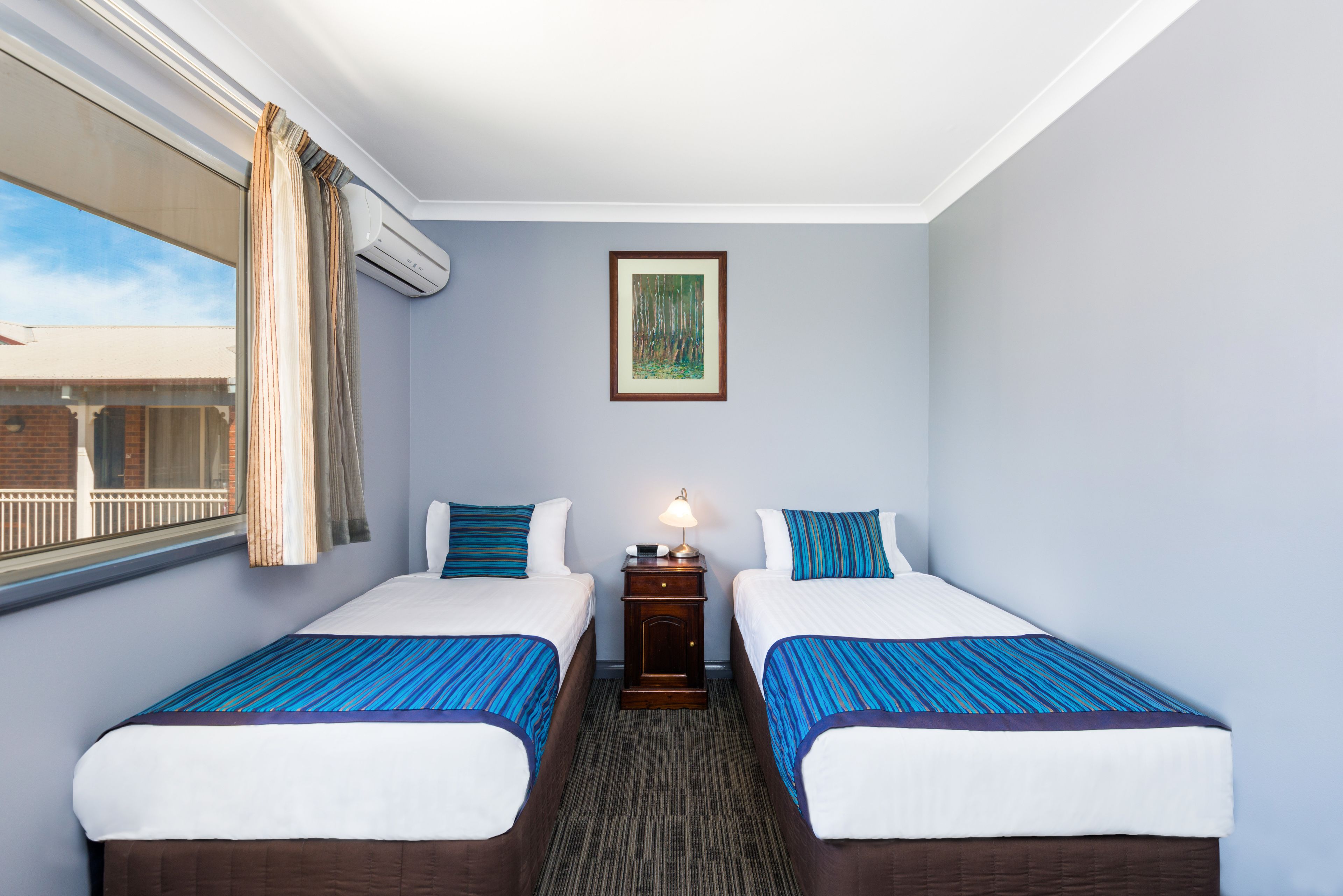 Une chambre d’hôtel avec deux lits, une table de chevet avec une lampe, un tableau encadré au mur et une fenêtre avec des rideaux.