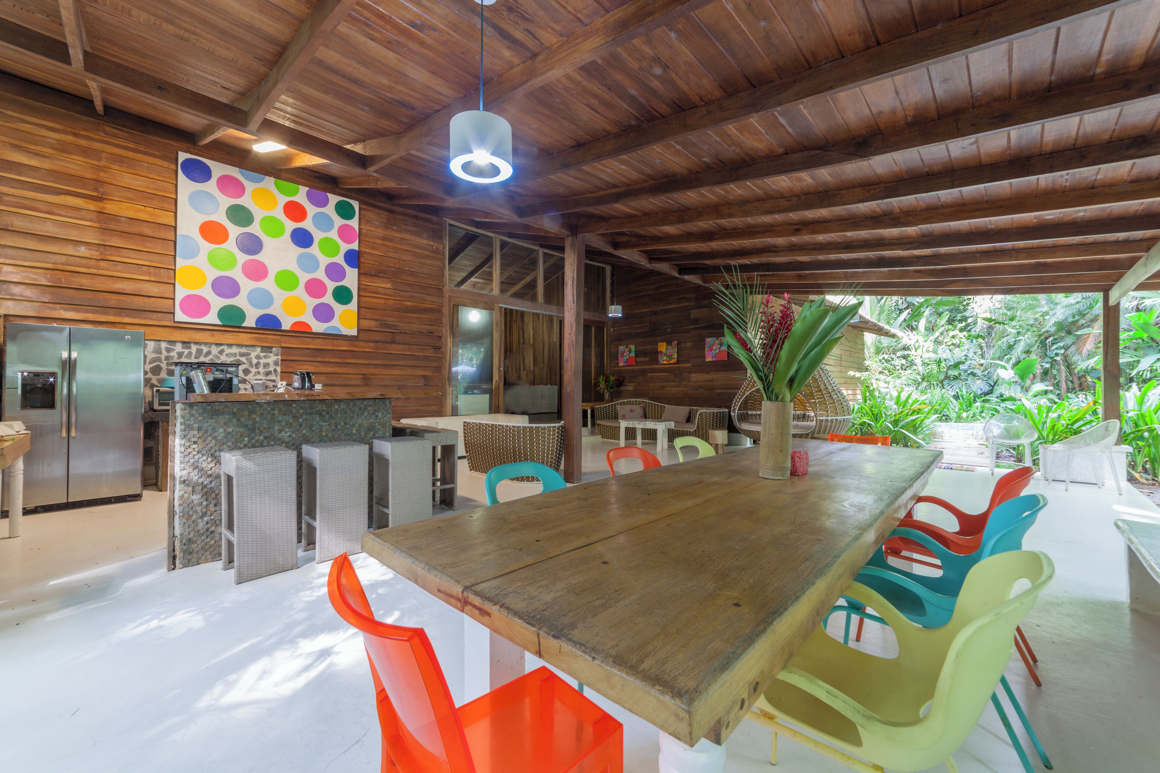 A spacious kitchen with a wooden ceiling, a large dining table with colorful chairs, and a view of lush greenery outside.
