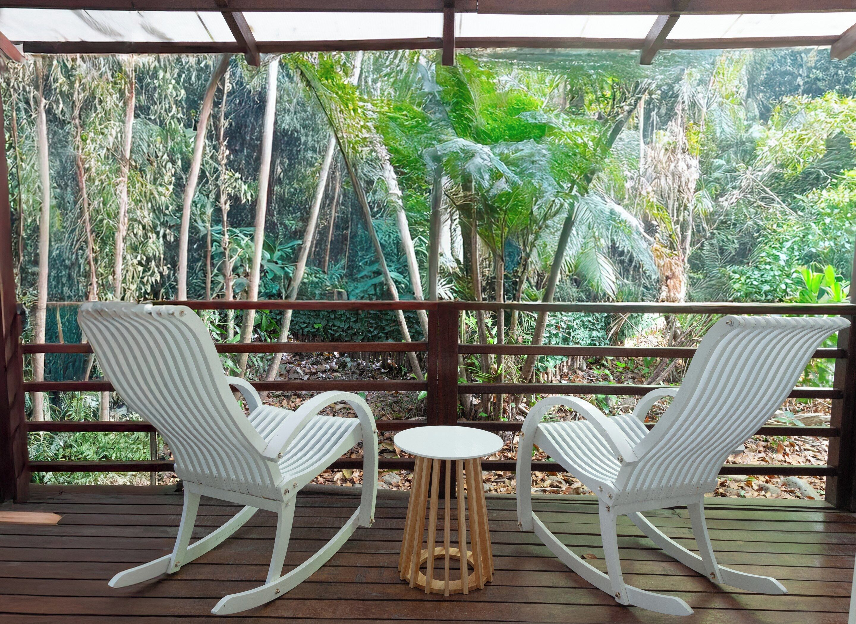 Una terraza de madera con dos mecedoras blancas y una pequeña mesa redonda, con vistas a un exuberante jardín tropical.