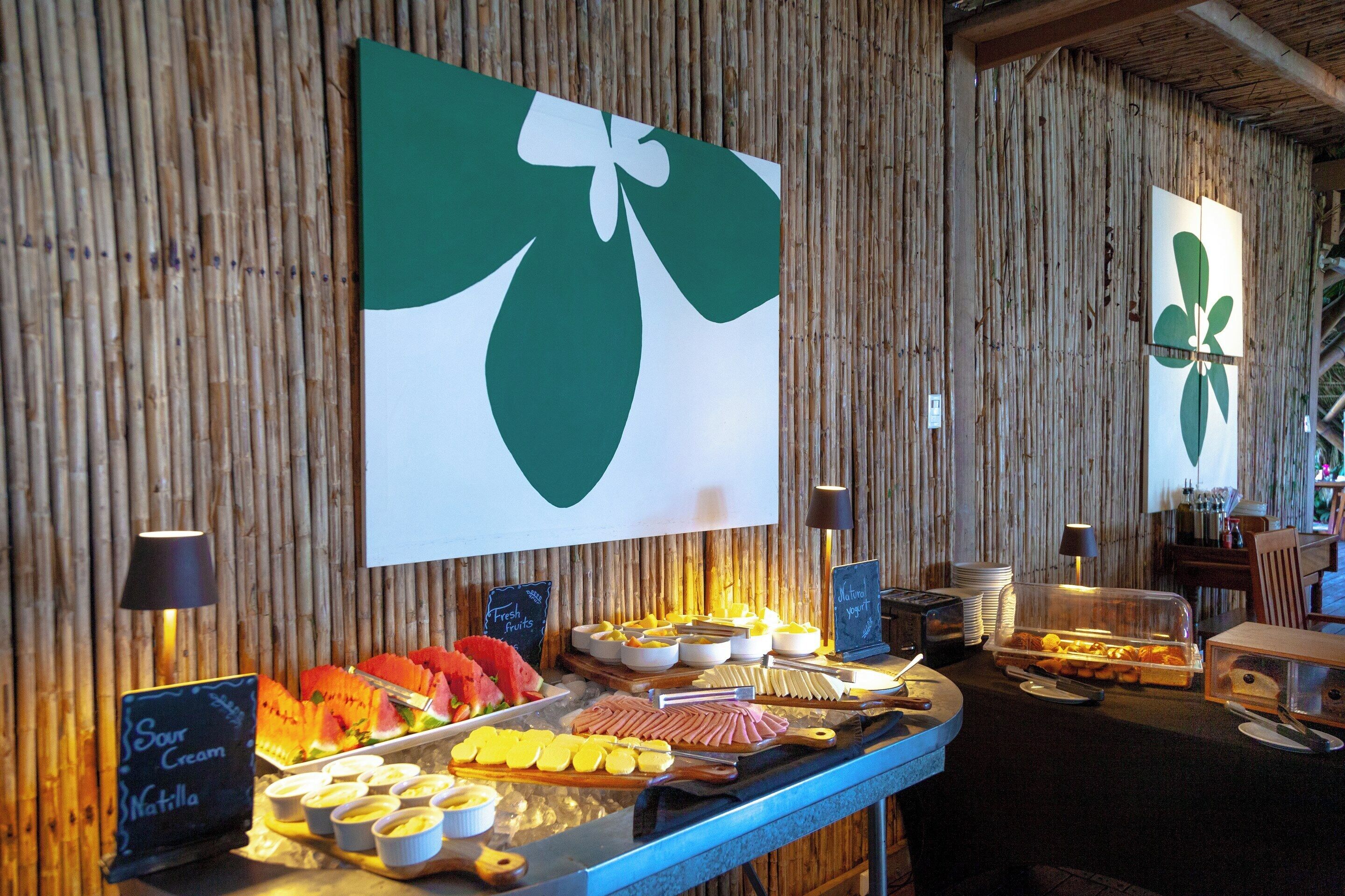 Un bufé con diversos alimentos, entre ellos frutas, quesos y carnes, con un fondo de bambú y una decoración de hoja verde grande.