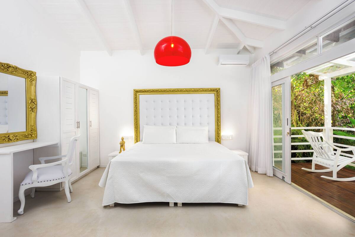 A bedroom with a large bed, a desk, and a chair. There is a balcony with a white rocking chair.