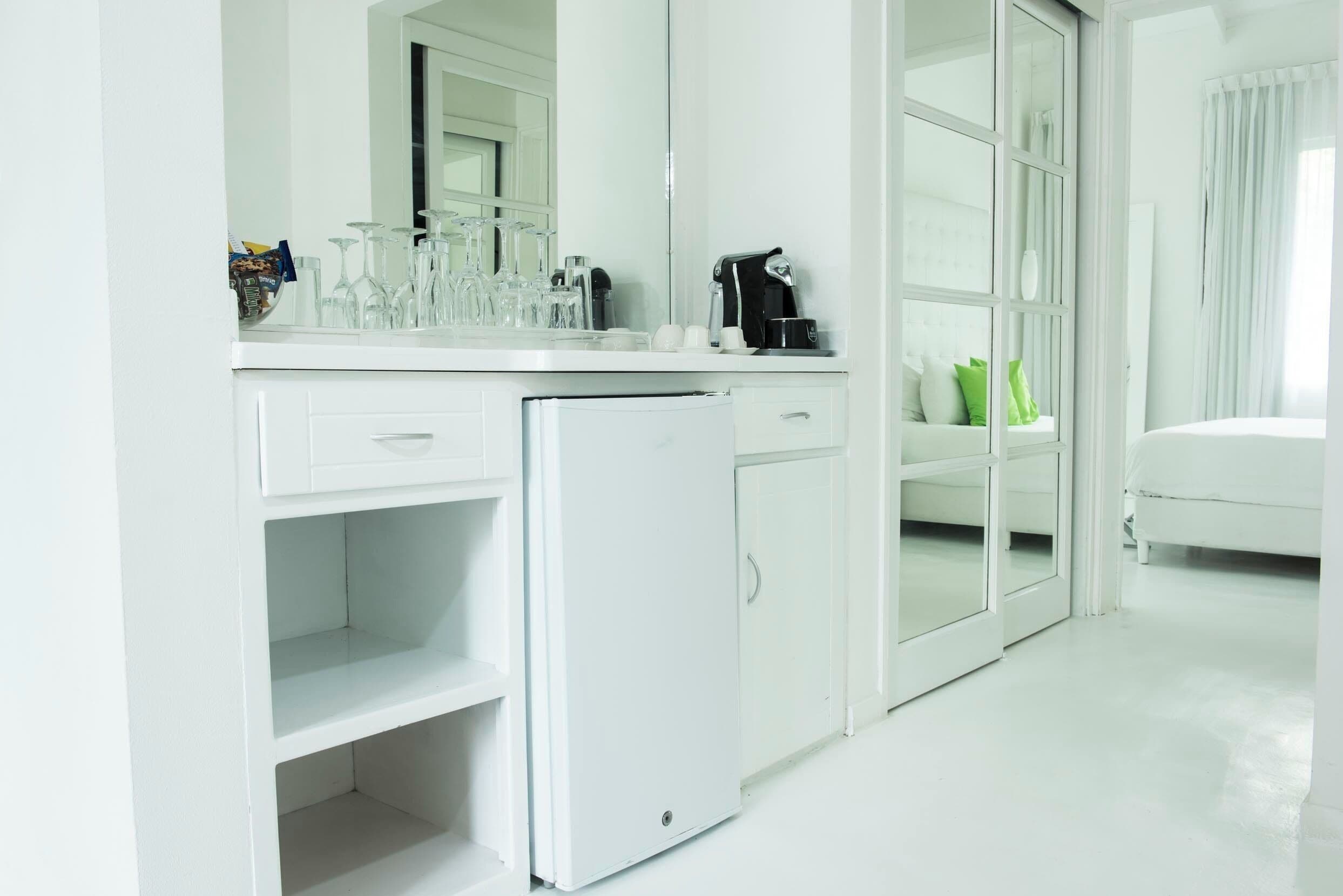 Habitació d'hotel moderna i blanca amb una cuineta compacta, un refrigerador petit i un llit amb almoades verdes.