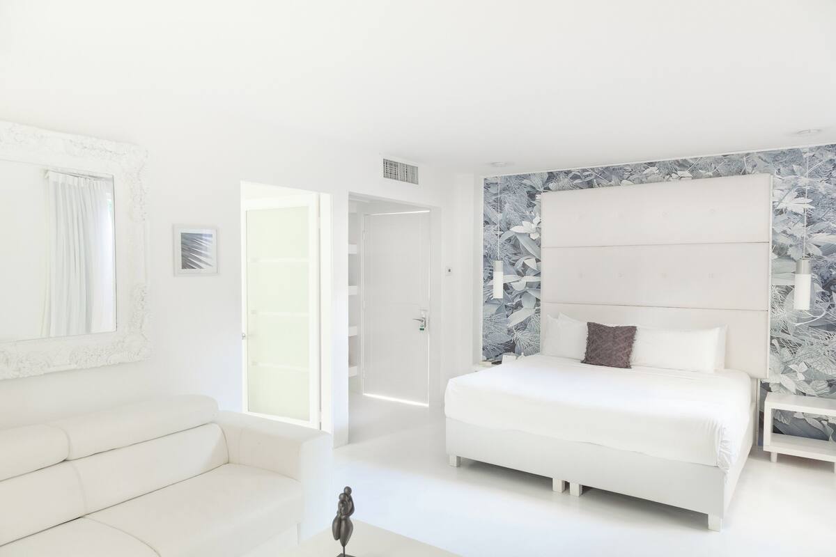 A modern bedroom with a large bed, a white sofa, and a door leading to another room.