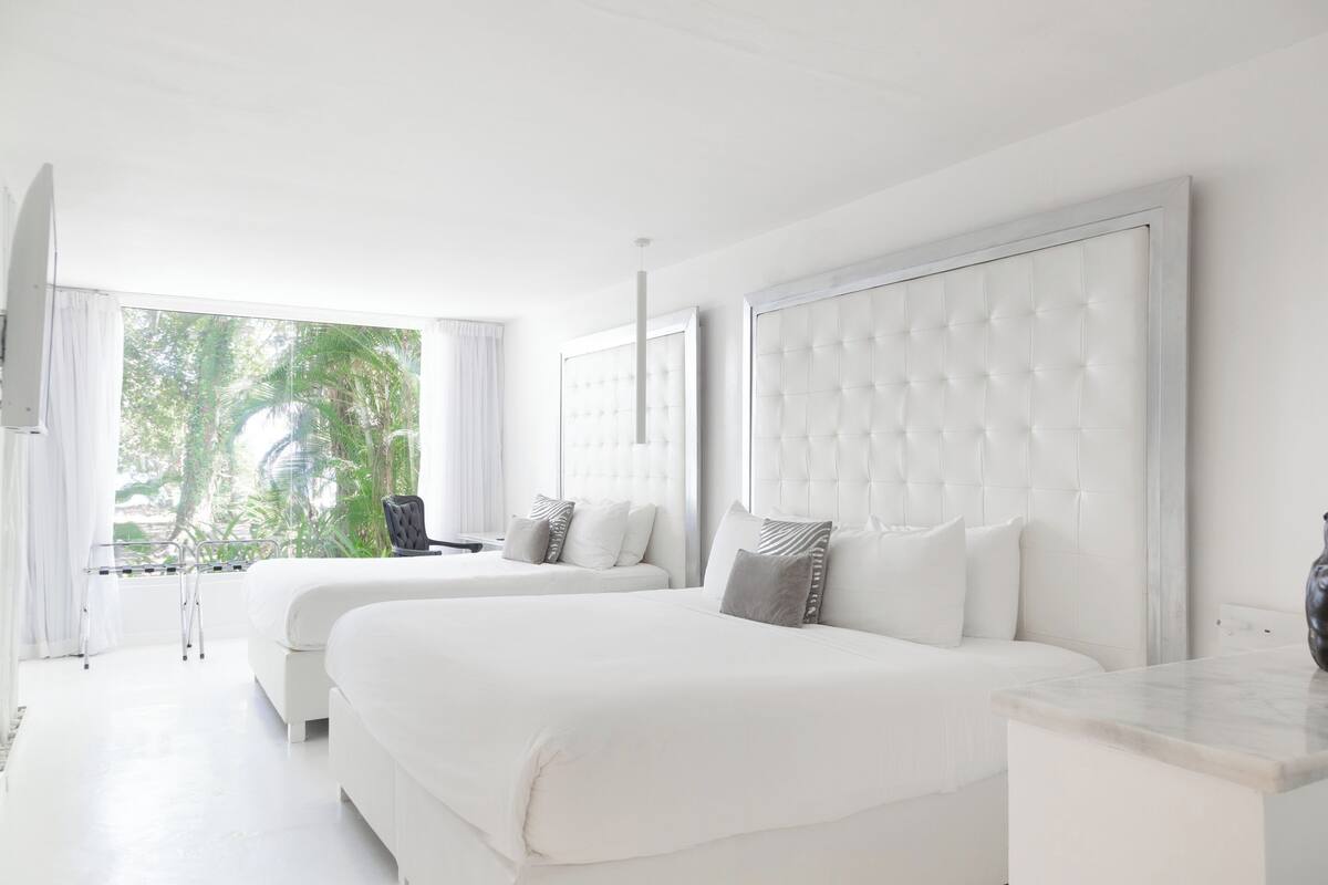 A modern hotel room with two large beds, a marble side table, and a view of greenery outside.
