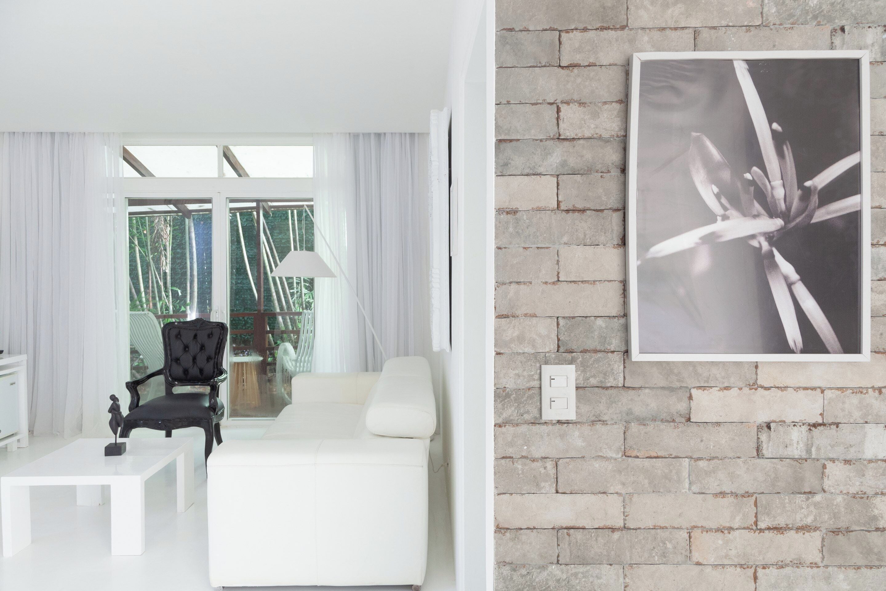 A modern living room with a white sofa, a black armchair, and a white coffee table. There is a large window with sheer curtains, a lamp on a side table, and a framed photograph on the brick wall.