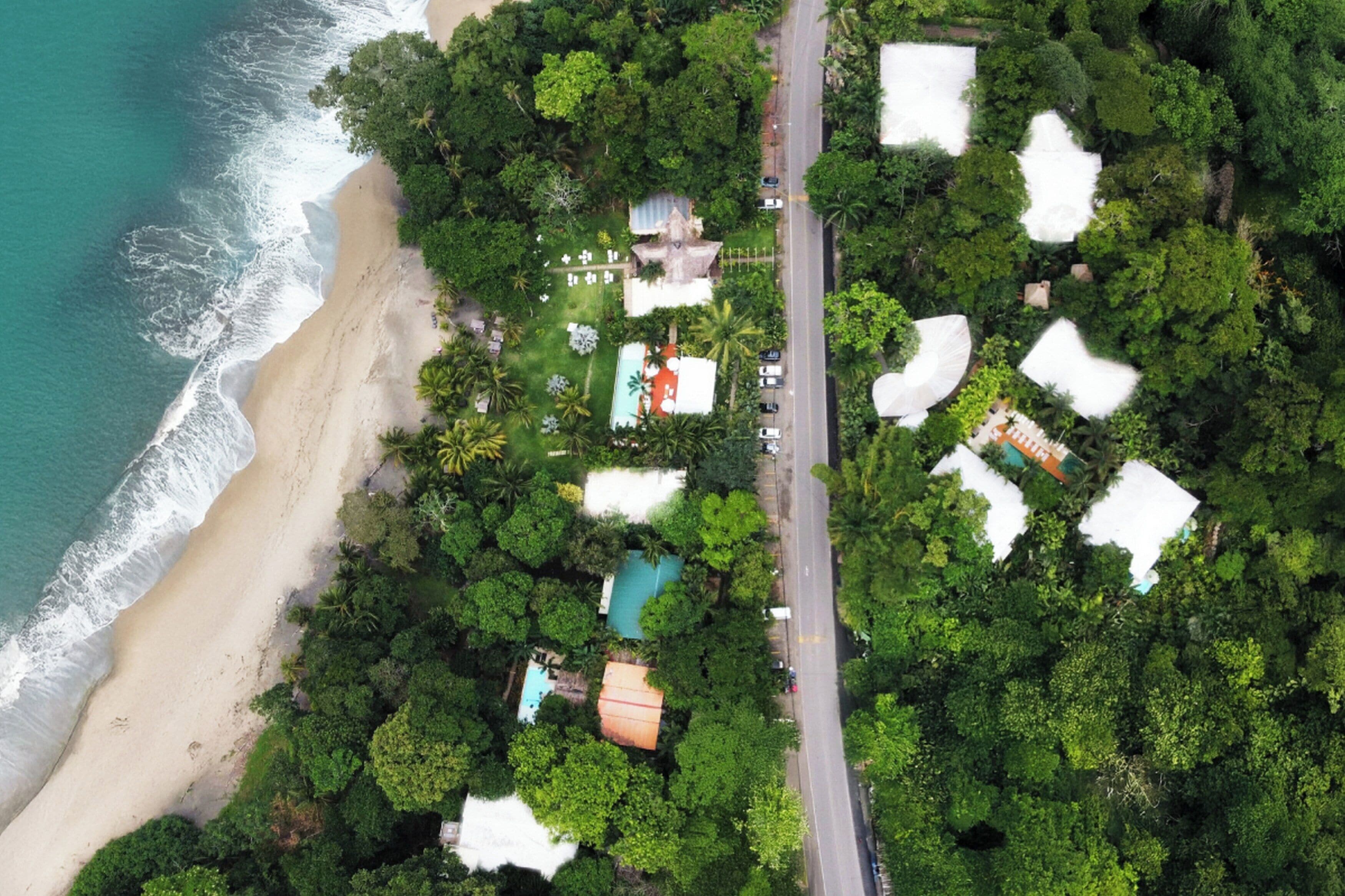 Vista aérea de una zona costera con una carretera, playa y varios edificios rodeados de vegetación densa.