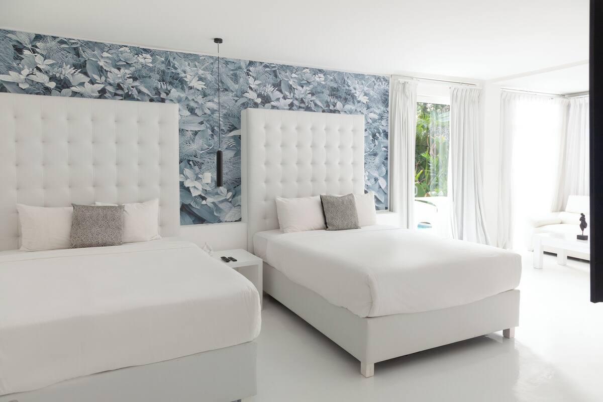 A modern bedroom with two beds, a large floral mural, and a view of greenery outside.