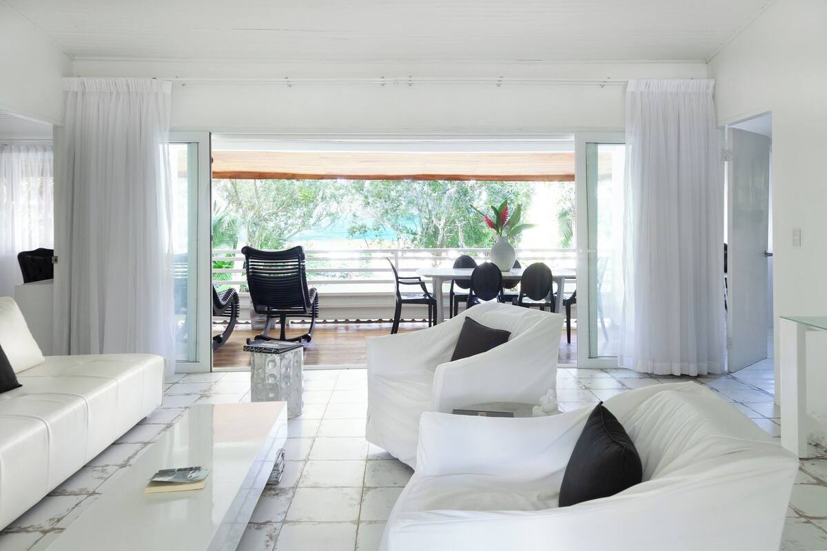 A modern living room with white sofas, a glass coffee table, and a view of a balcony with outdoor furniture.
