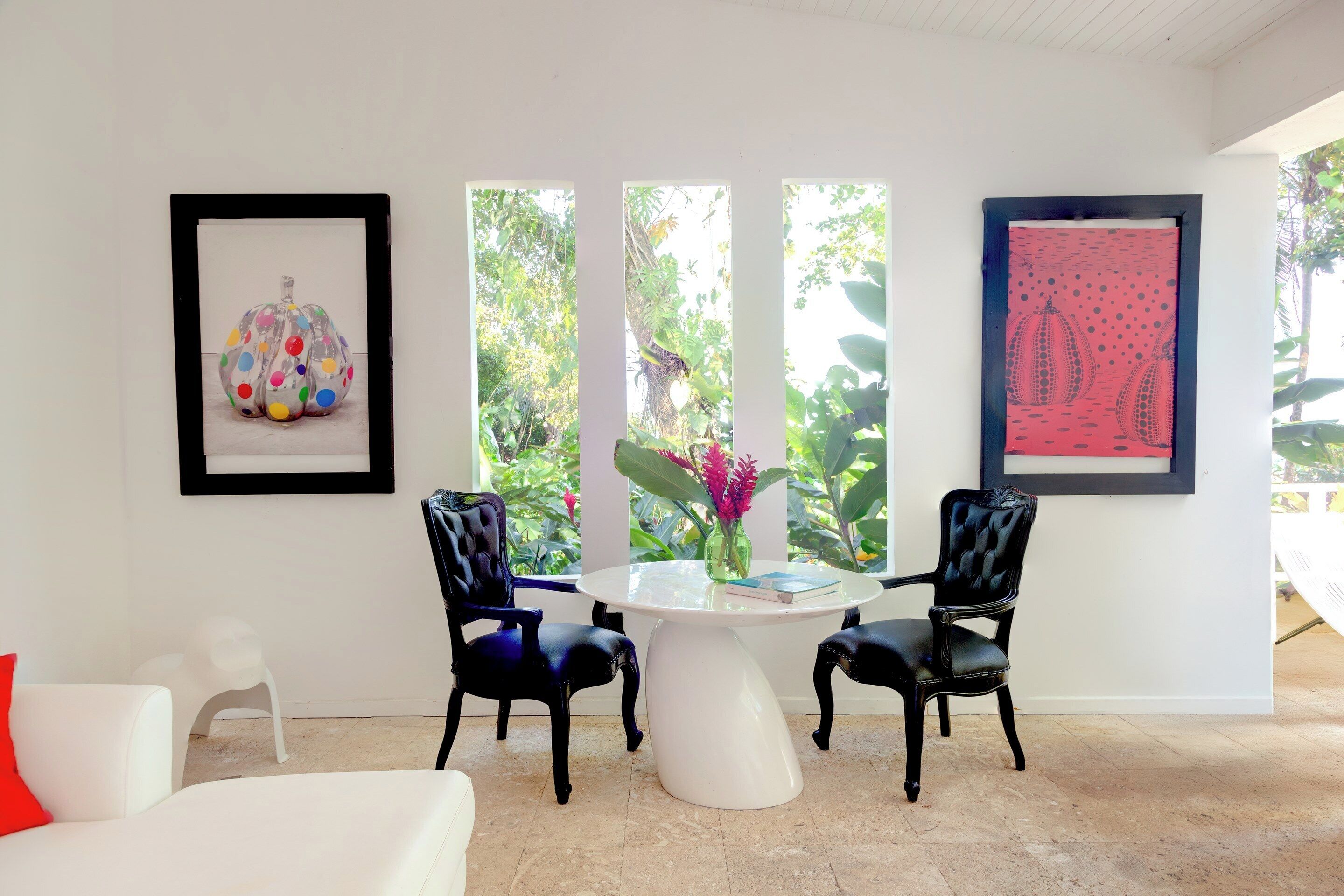 Une pièce avec une table ronde blanche, deux chaises noires, un canapé blanc et un coussin rouge. Les murs sont ornés d’œuvres d’art encadrées et une fenêtre offre une vue sur la verdure.