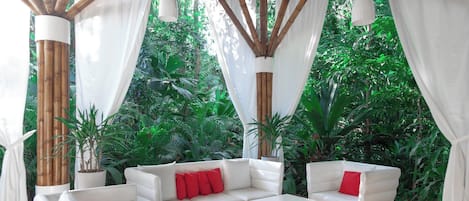 A covered outdoor seating area with white furniture and bamboo pillars, surrounded by lush greenery.