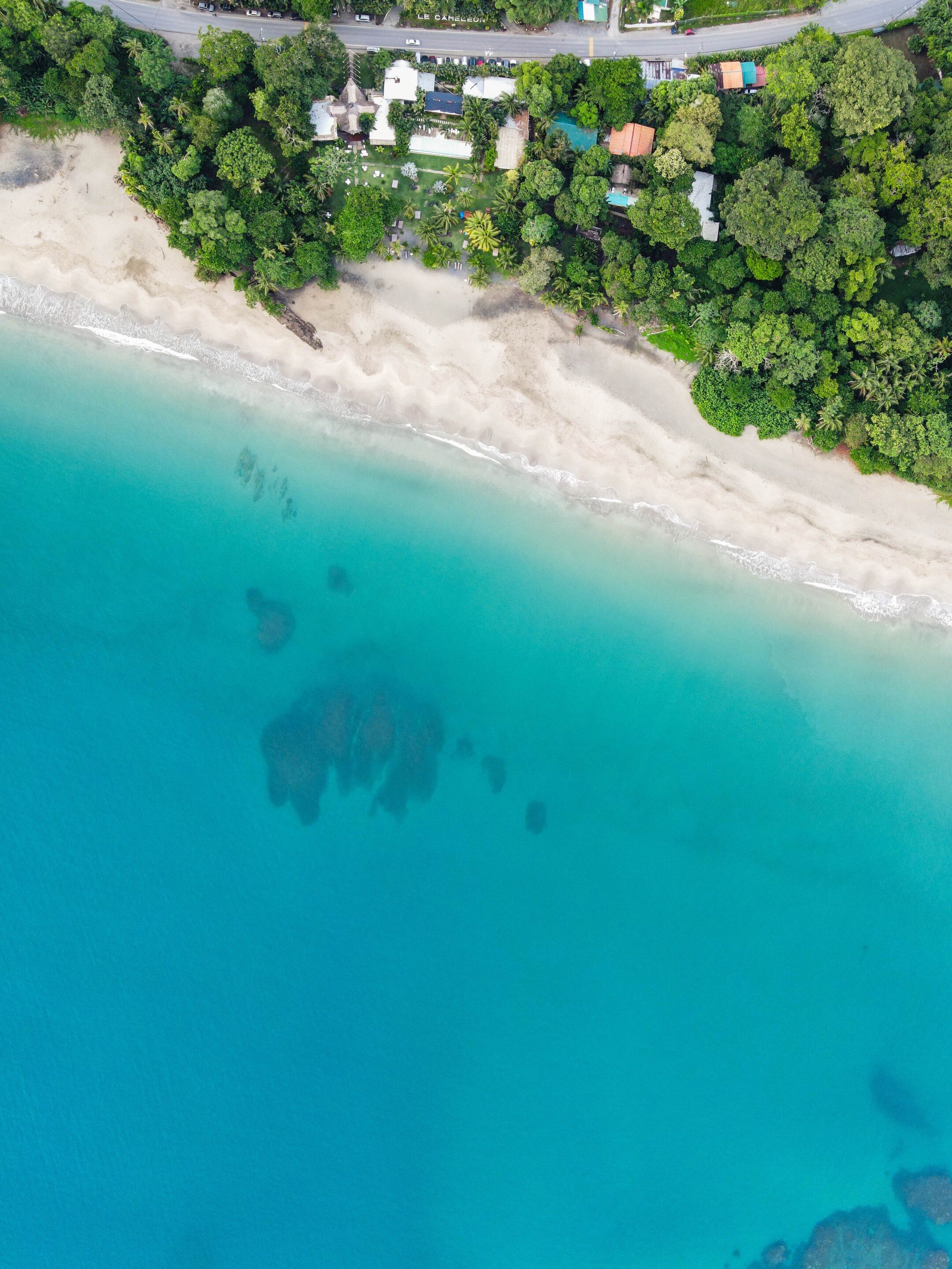 An aerial view of a beach with clear turquoise waters, a sandy shore, and a cluster of buildings with a mix of architectural styles.
