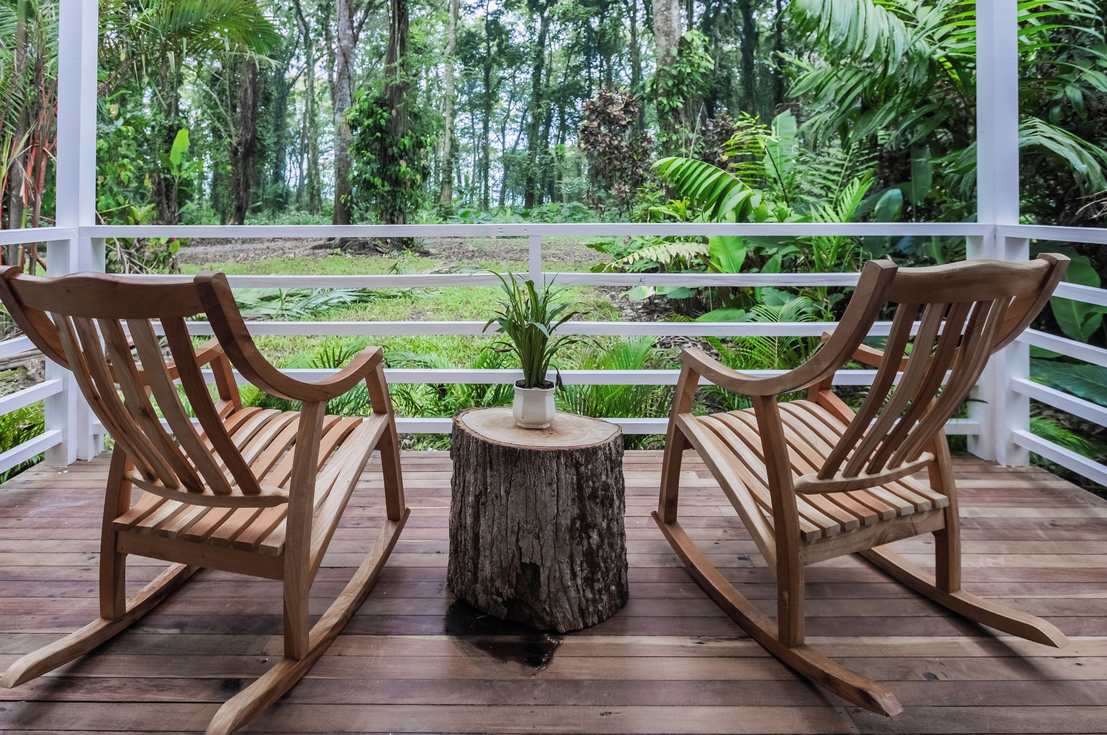 Two wooden rocking chairs on a wooden deck with a white railing, overlooking a lush green forest.