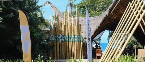 Resort à beira-mar com estrutura de bambu, placa indicativa da praia e vegetação exuberante.