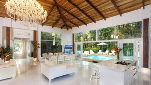 A spacious living area with a white sofa, a dining table, and a kitchen island with a built-in stove. Large windows provide views of a swimming pool and lush greenery. - Le Cameleon Puerto Viejo (Cahuita)