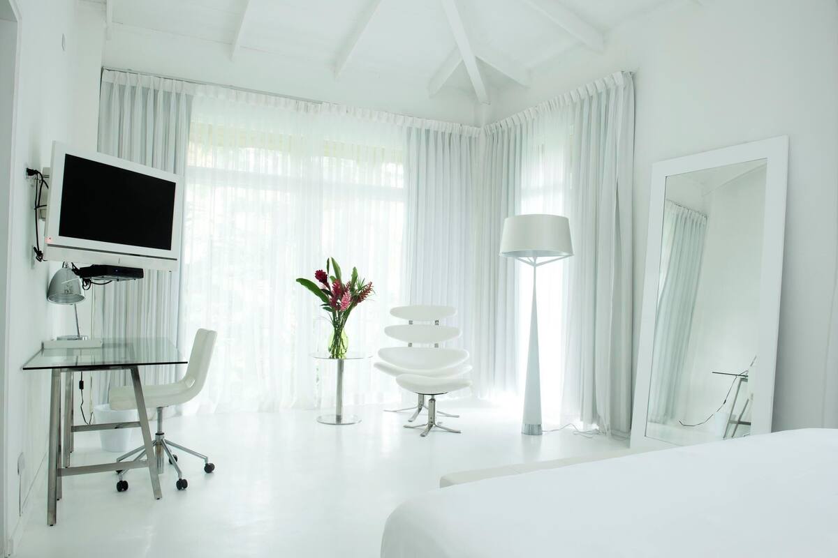 A modern hotel room with a large bed, a desk with a computer, a chair, a lamp, and a mirror.