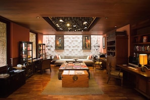 A cozy hotel room with a wooden coffee table, a sofa, a bookshelf, and a desk with a lamp.