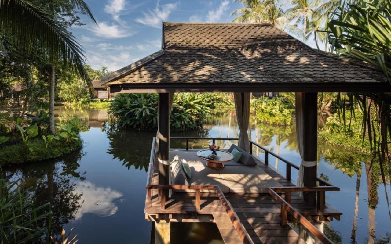 A wooden deck with a thatched roof structure, surrounded by water and greenery.