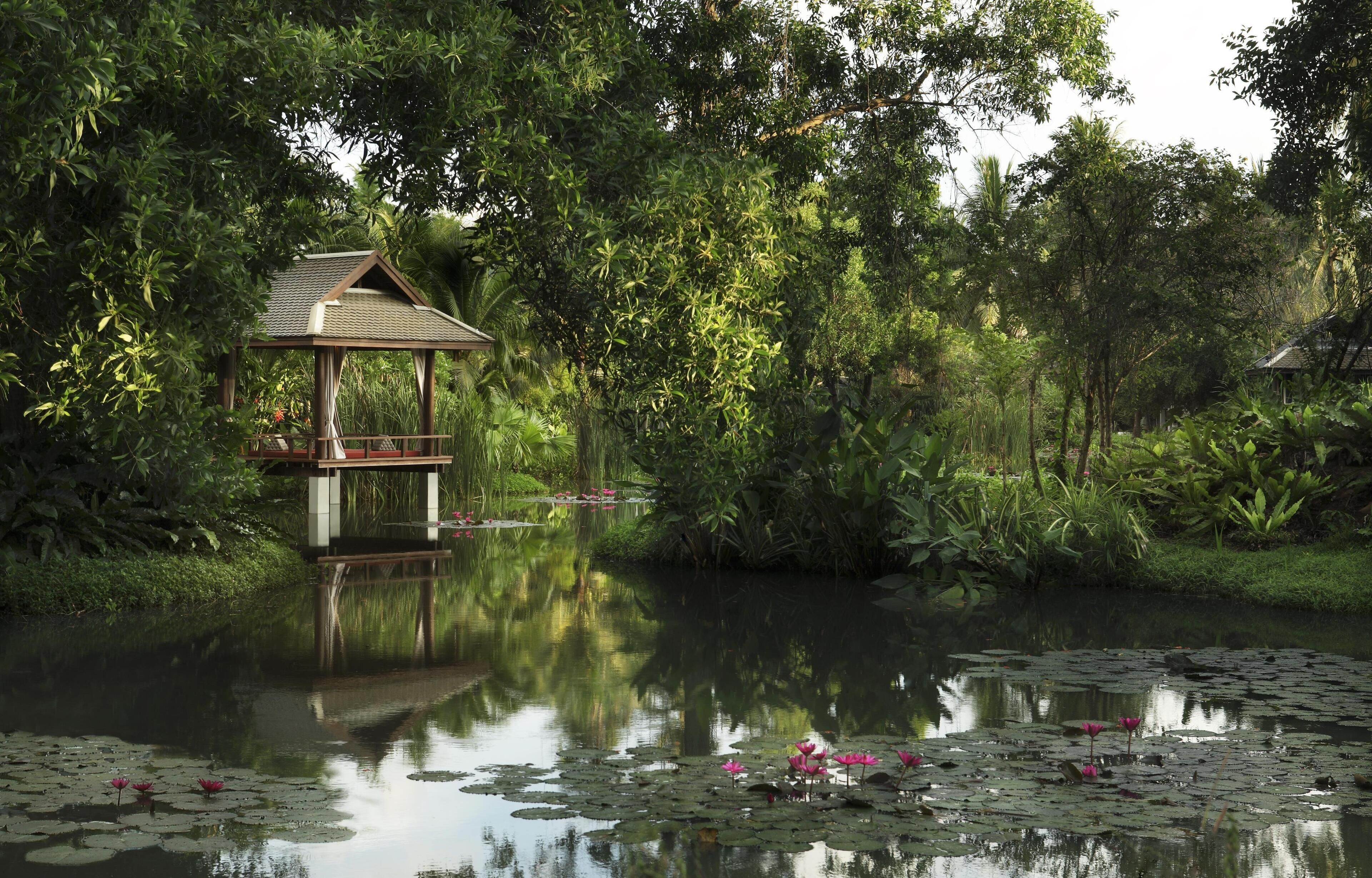 A tranquil pond with lily pads and pink flowers, surrounded by lush greenery and a wooden gazebo.