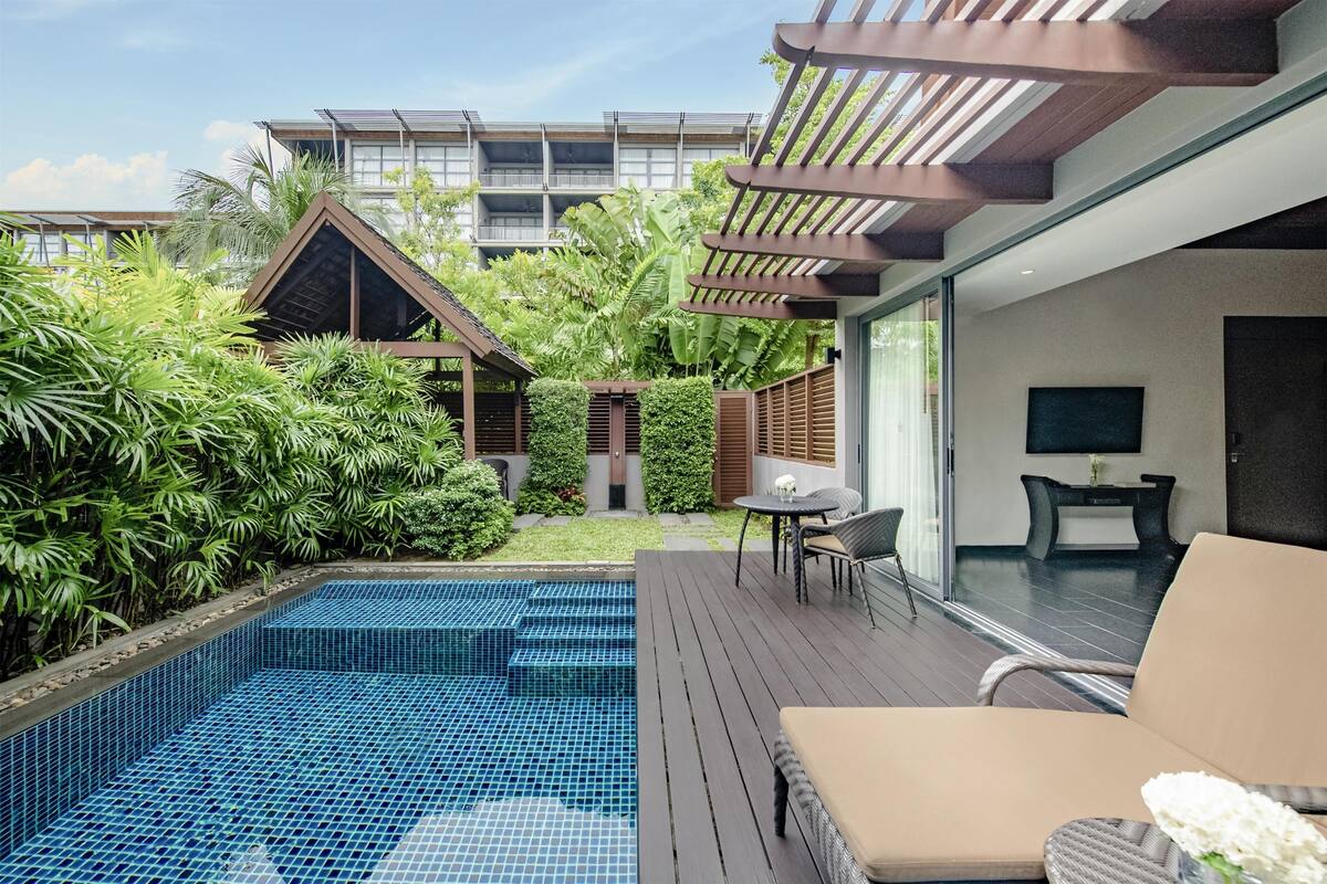 Ein moderner Poolbereich im Freien mit Holzterrasse, einer Sitzecke mit Tisch und Stühlen sowie üppiger Bepflanzung.
