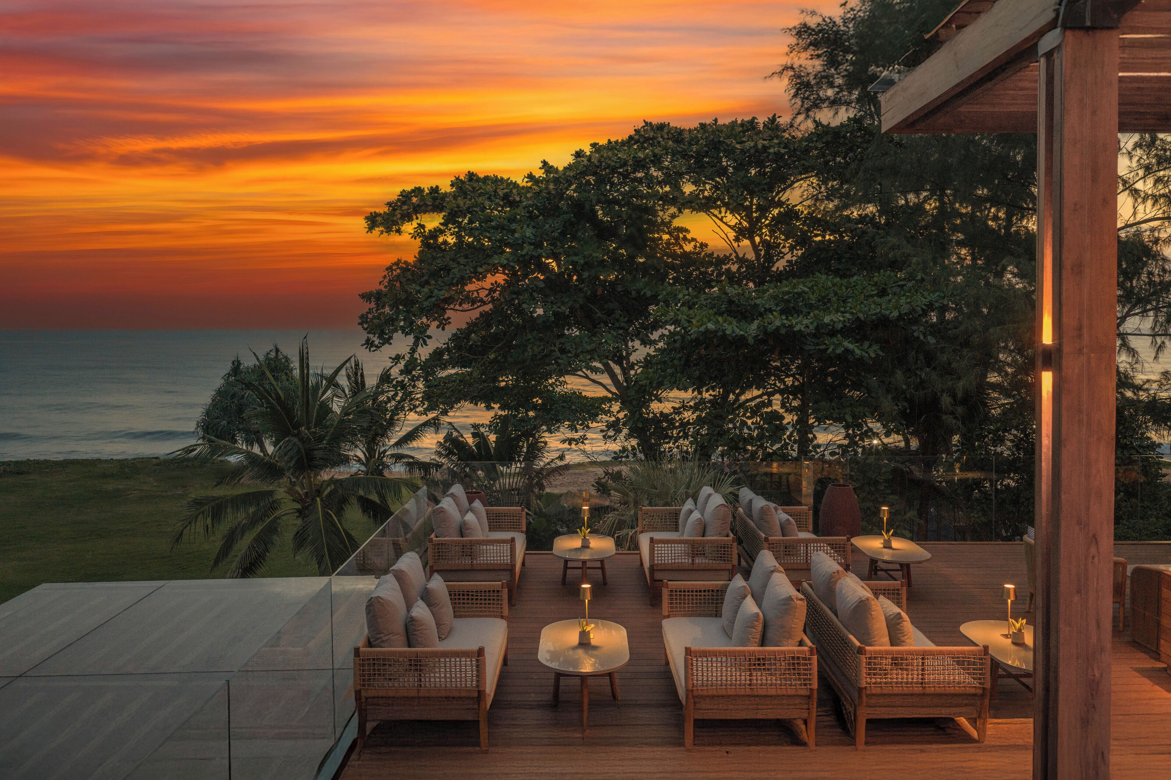 A deck with wicker furniture and tables, overlooking a sunset and ocean view.