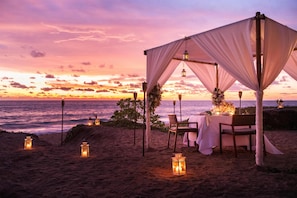 A beachside dining setup with a canopy, tables, chairs, and lanterns.