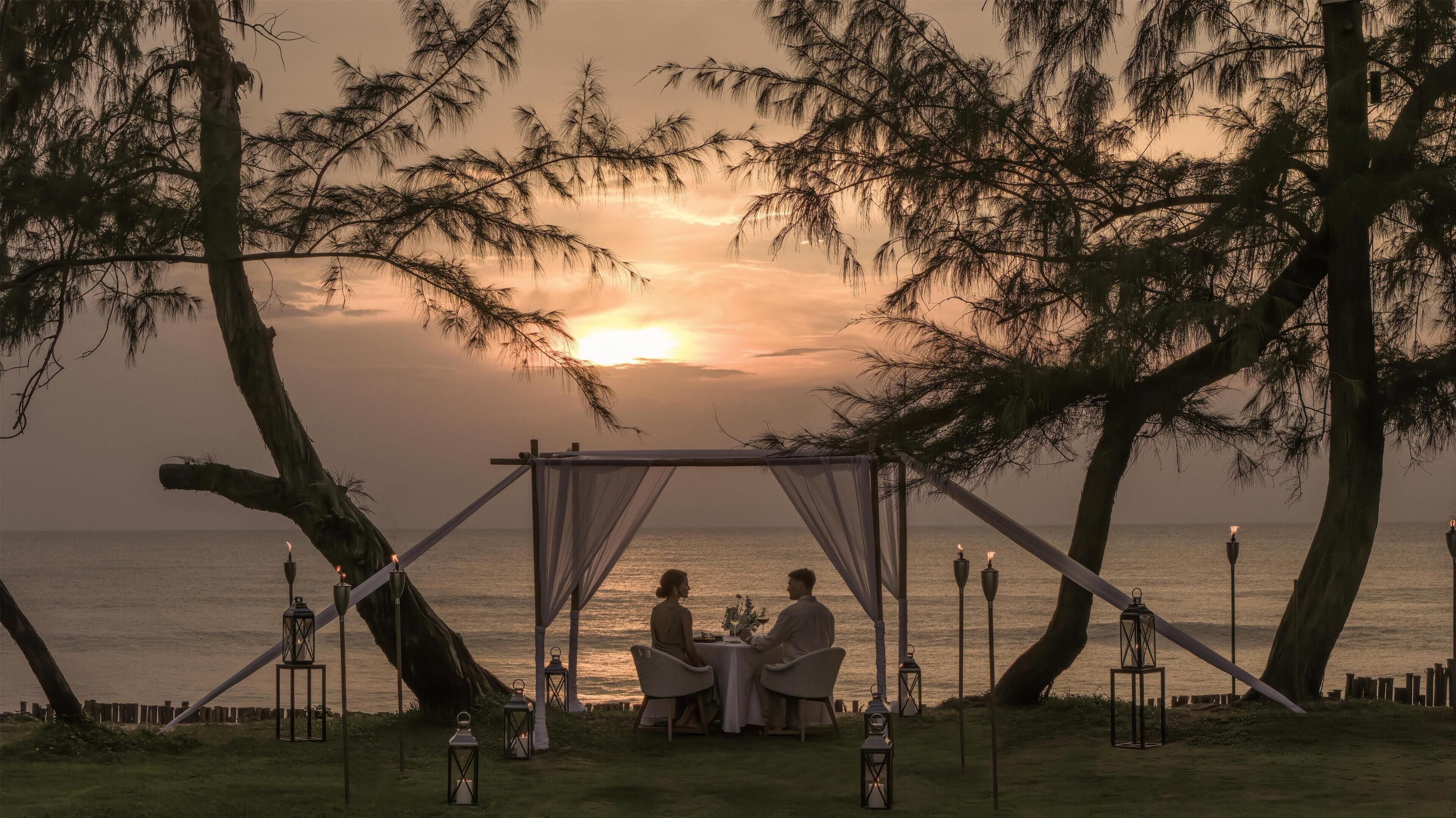 A couple dining under a canopy at sunset by the sea.
