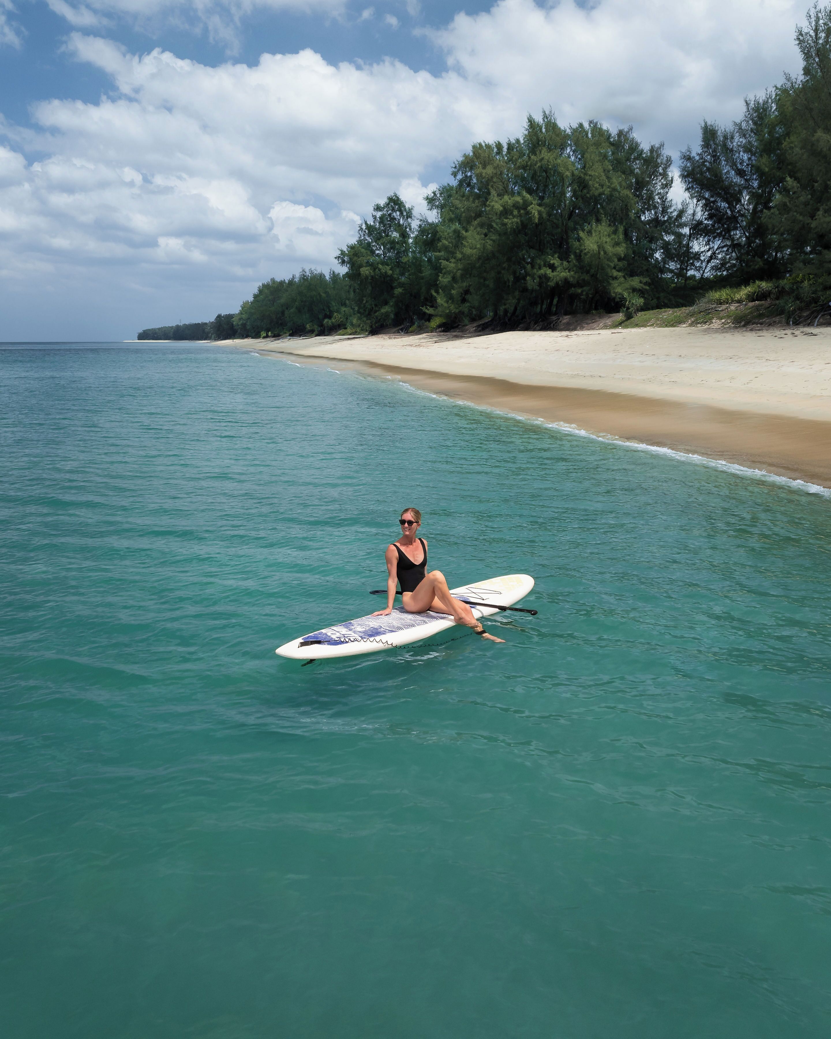 Une personne est assise sur une planche à pagaie dans une eau turquoise et limpide, avec une plage de sable et des arbres en arrière-plan.