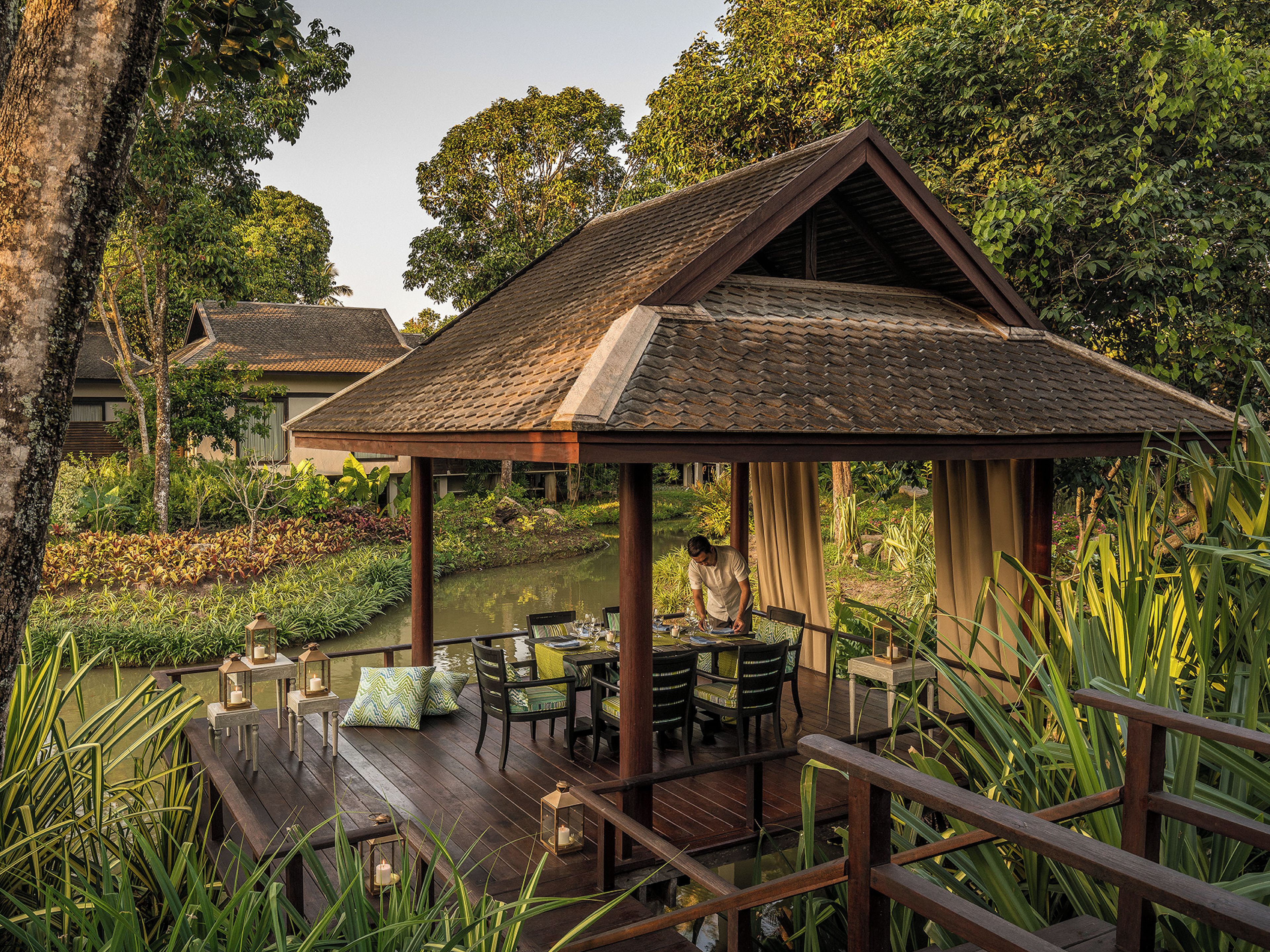 A wooden deck with tables and chairs, surrounded by lush greenery and a calm water body.