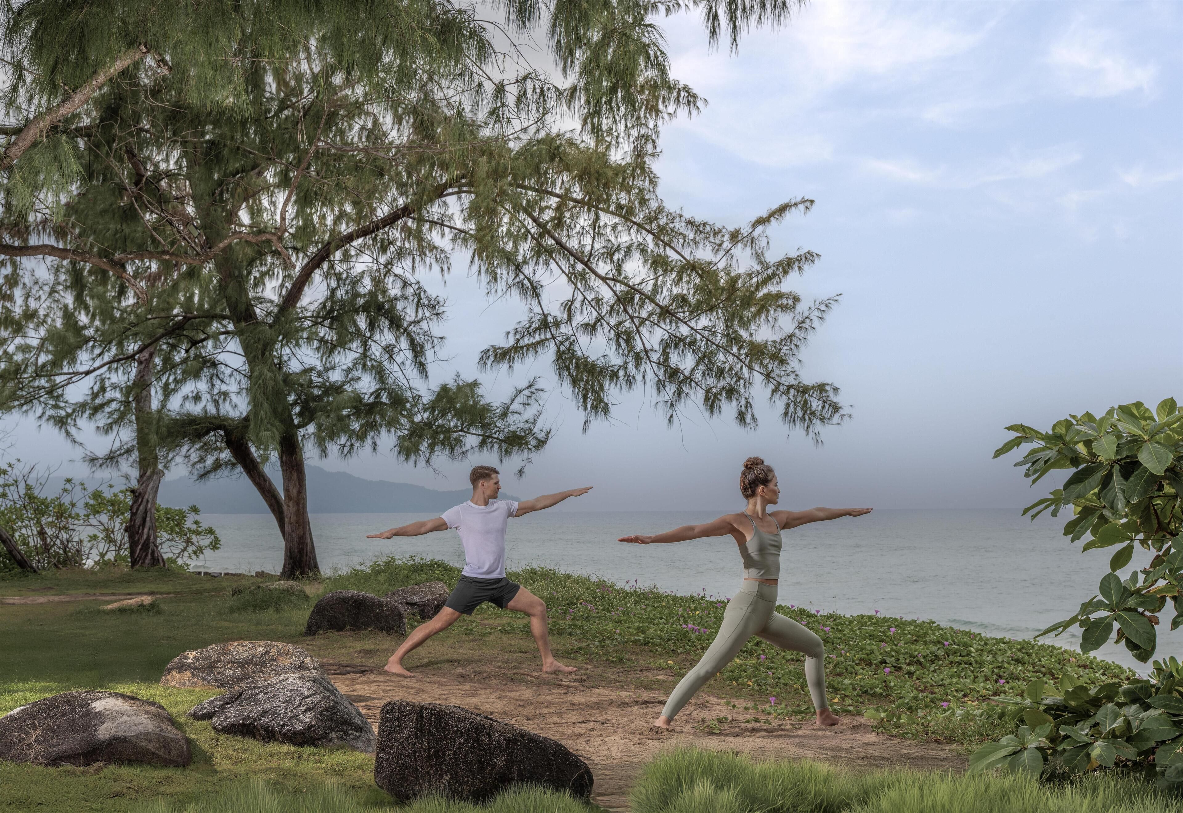 Duas pessoas praticando yoga ao ar livre perto de um corpo d'água, com árvores e pedras.
