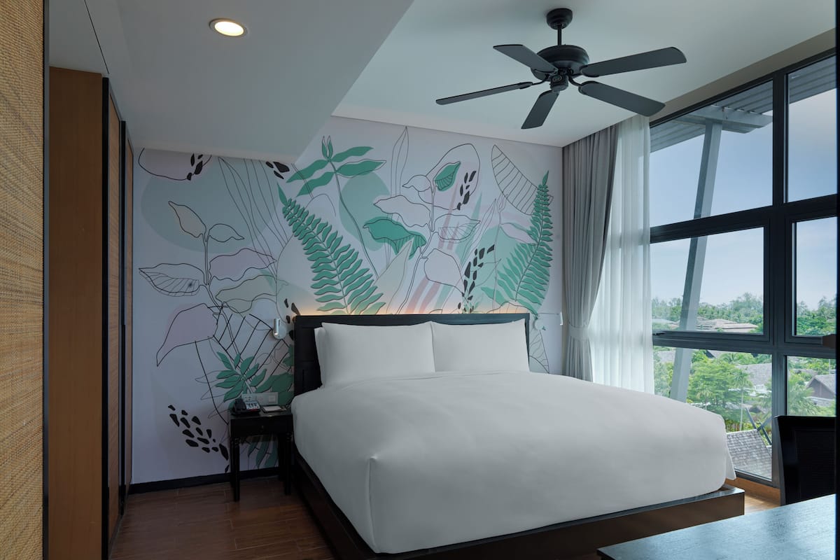 Ein modernes Hotelzimmer mit einem großen Bett, einem Deckenventilator und Blick ins Freie durch große Fenster.