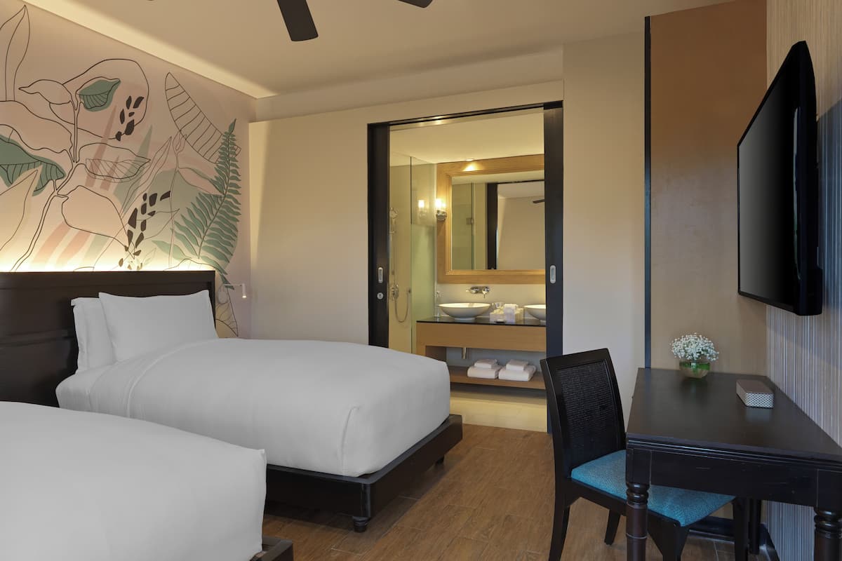 Ein Hotelzimmer mit zwei Betten, einem Schreibtisch, einem Stuhl, einem Fernseher und einem Deckenventilator. Ein Badezimmer ist durch eine geöffnete Tür zu sehen.