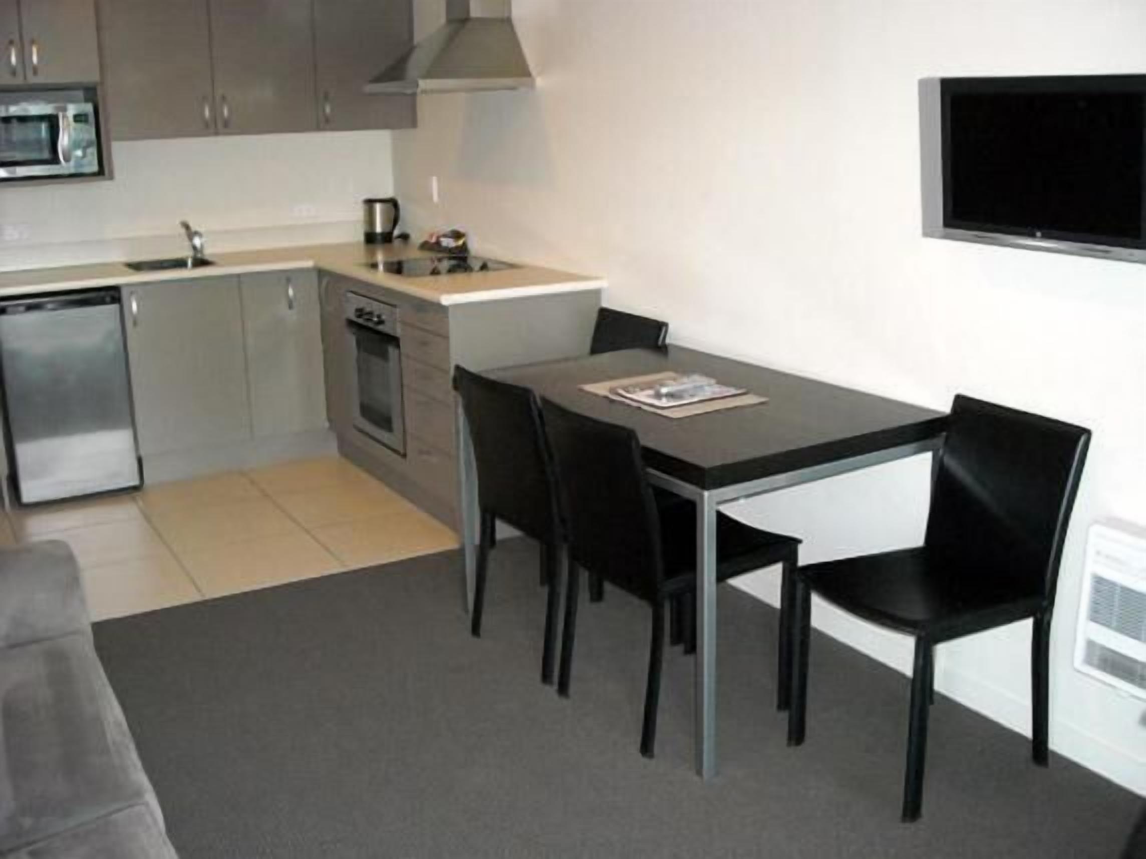 A compact kitchen with a microwave, stove, and sink. There is a dining table with four chairs and a flat-screen TV mounted on the wall.