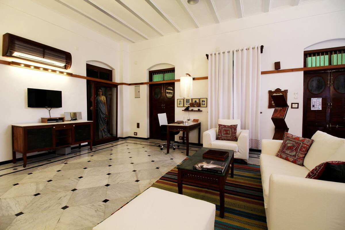 A spacious living room with a large sofa, a coffee table, and a television on a stand. There is a patterned rug on the floor and a wooden cabinet with a flat-screen TV.