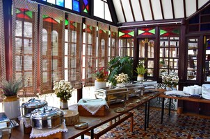 A dining area with buffet setup, large windows with patterned curtains, and potted plants.