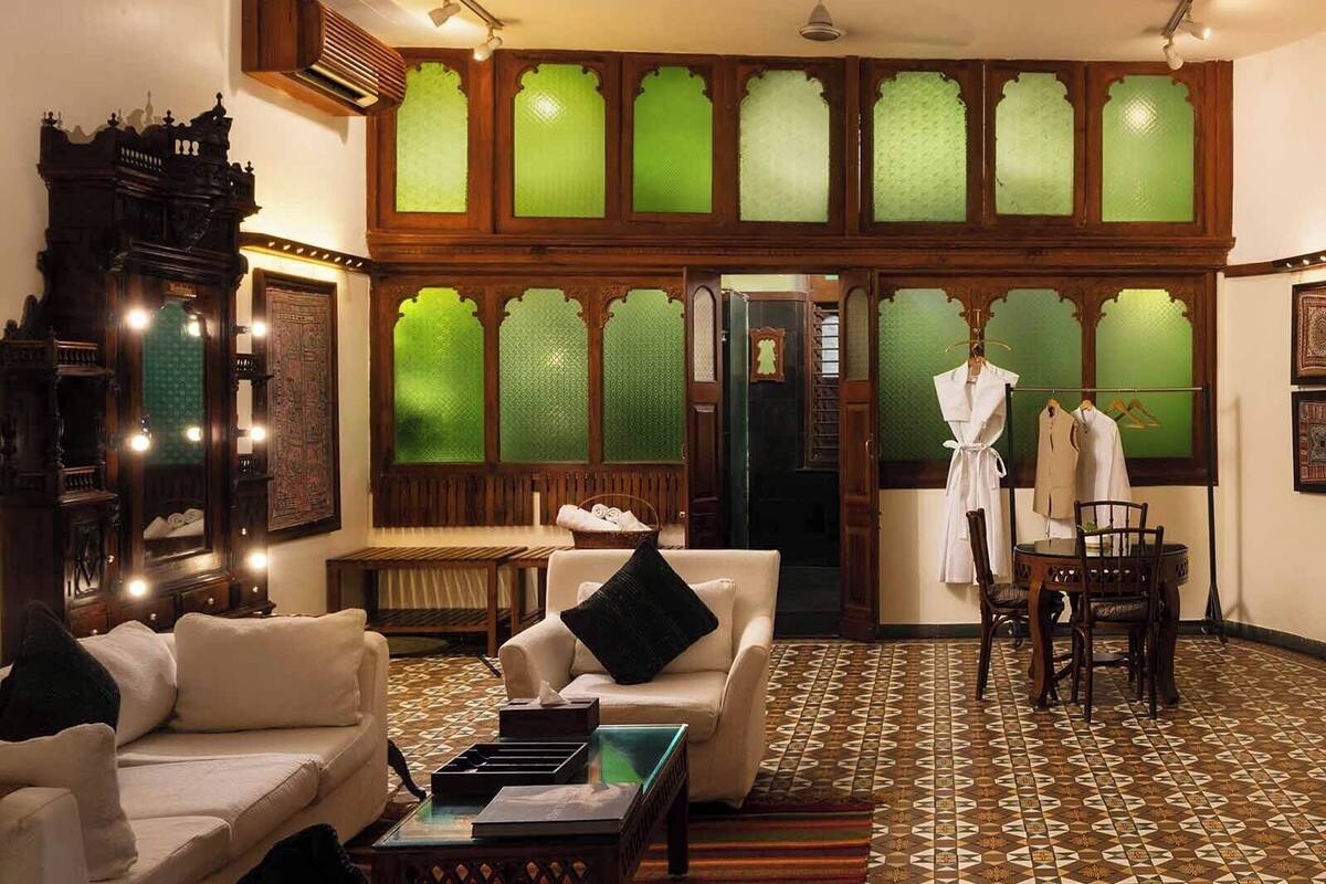A traditional room with a large mirror, a sofa, a small table, and a chair. There is a window with green glass panes and a patterned floor.