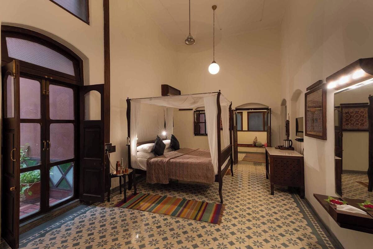 A bedroom with a four-poster bed, a small table with a lamp, a mirror, and a window with a view of the outside.