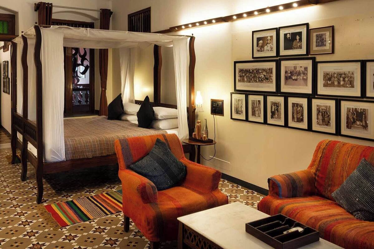 A four-poster bed with a canopy, a patterned rug, two armchairs, a small table, and a wall with framed photographs.