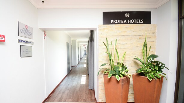 Een hotel lobby met een bord waarop 'Protea Hotels' staat, een gang met deuren en twee grote potplanten.