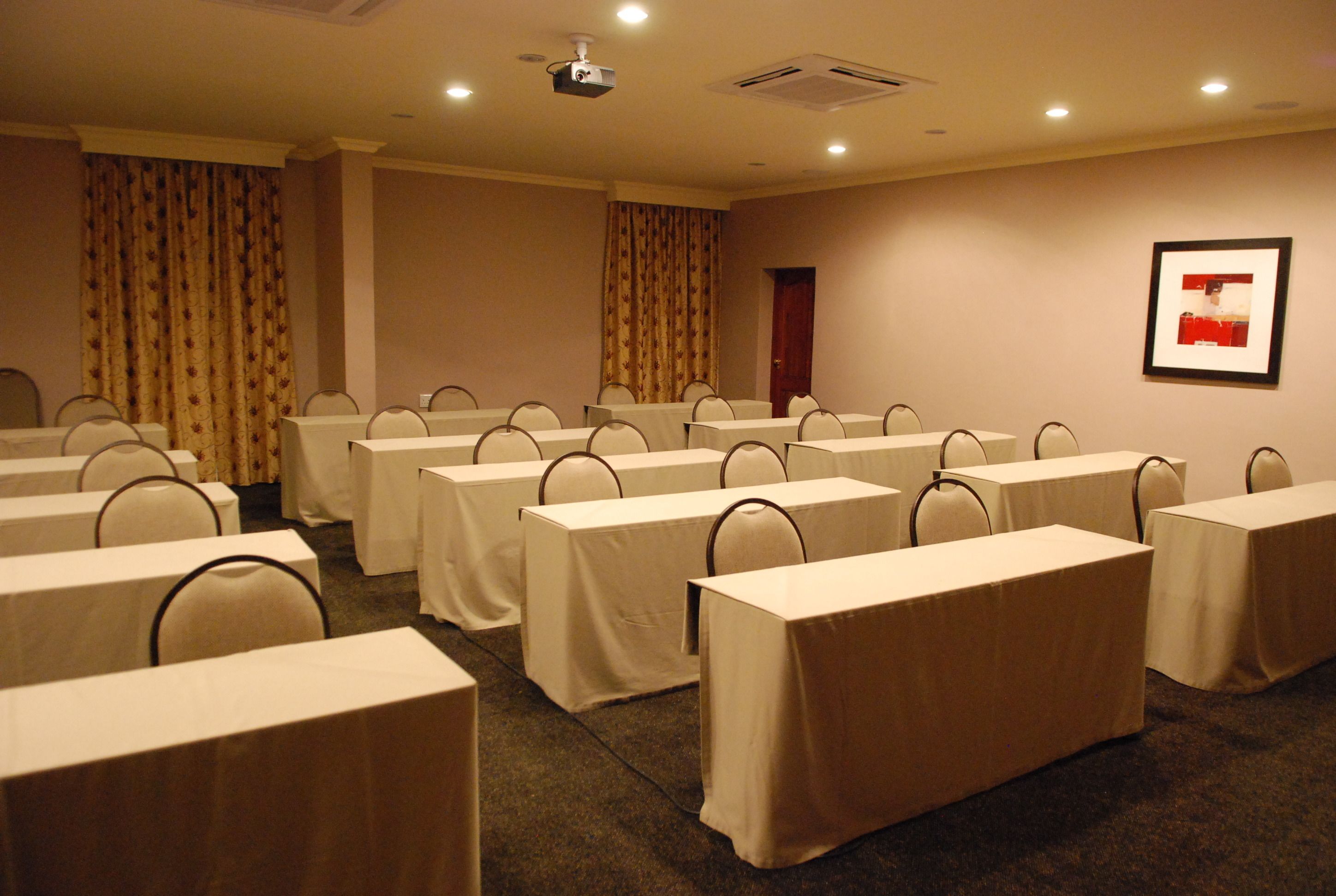 Un salón de conferencias con varias mesas y sillas, un proyector y un cuadro en la pared.