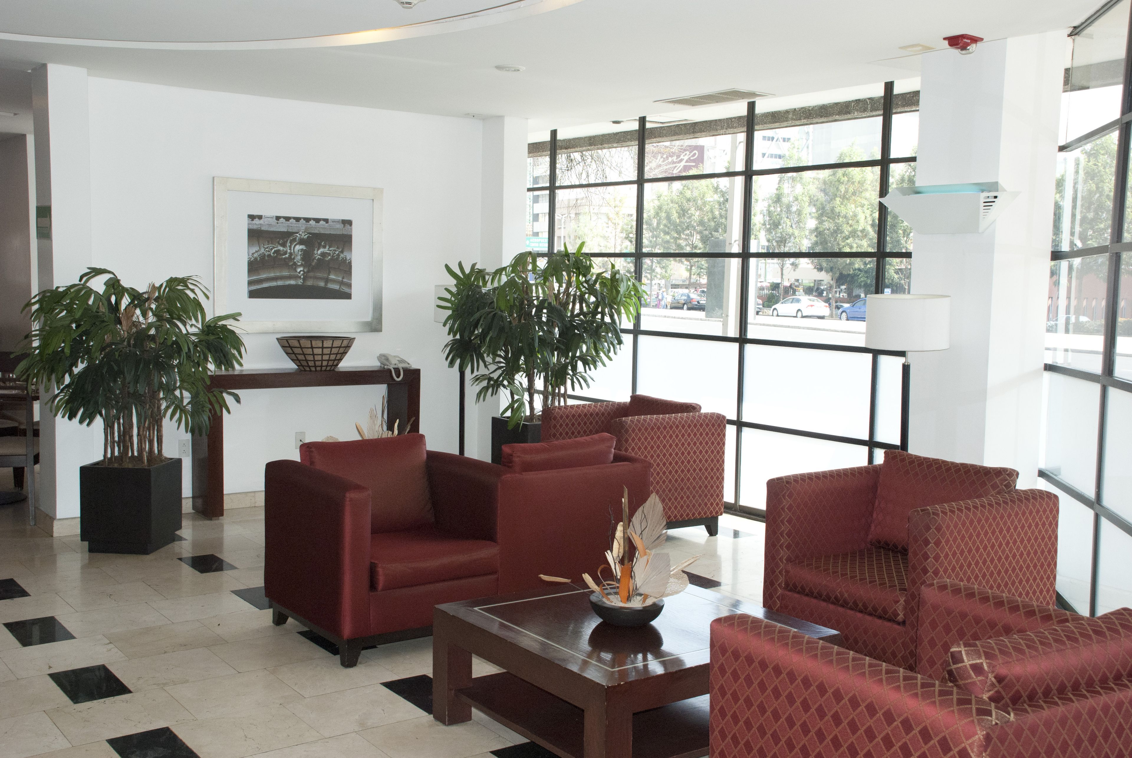 A lobby with red upholstered chairs, a coffee table with a decorative bowl, potted plants, and large windows with a view of the street.