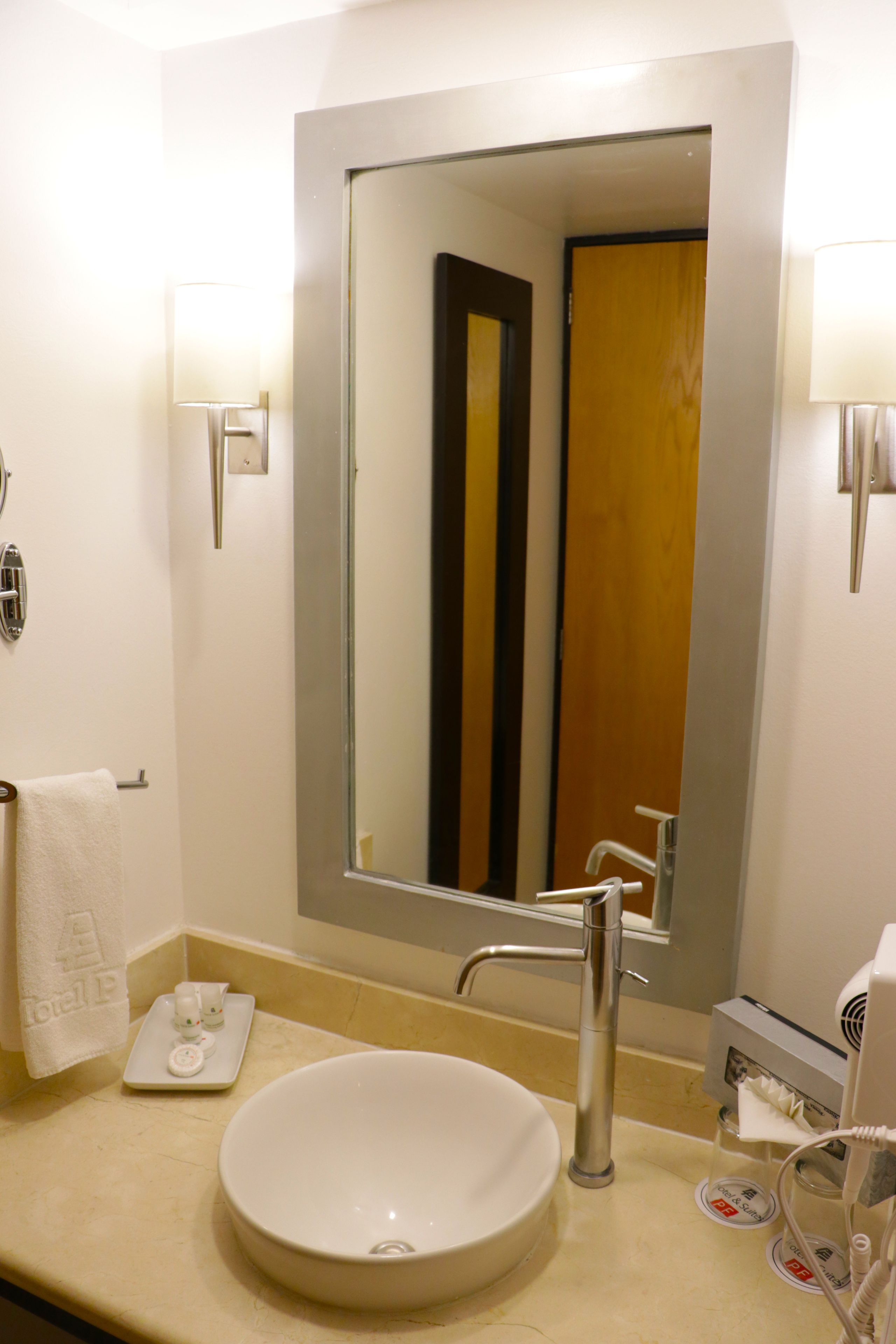 A bathroom with a large mirror, a round sink, a towel rack, and a soap dispenser.