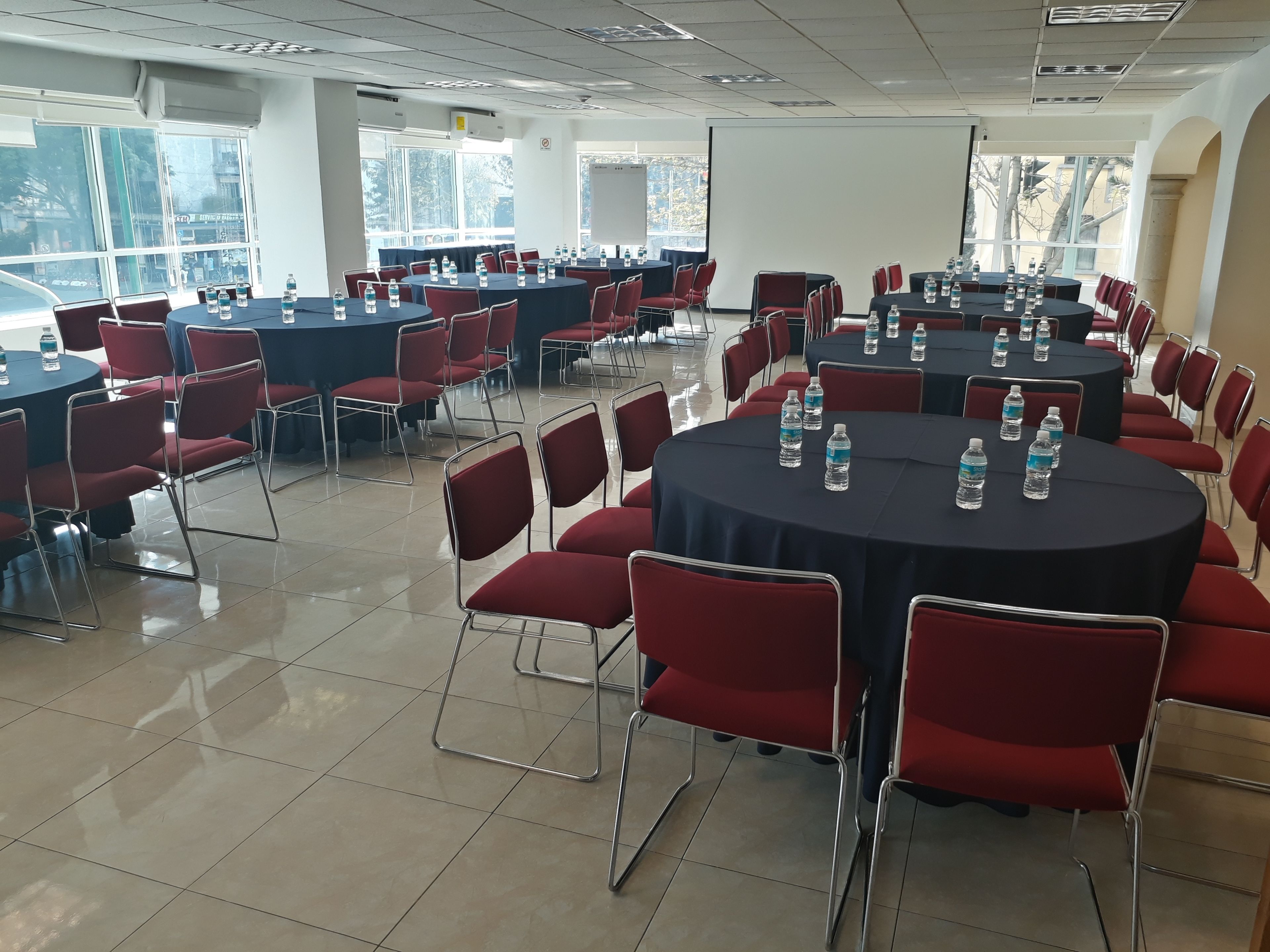 A conference room with multiple tables and red chairs, arranged in a U-shape. Tables are covered with black tablecloths, and each has several water bottles. The room has large windows allowing natural light, and the floor is tiled.