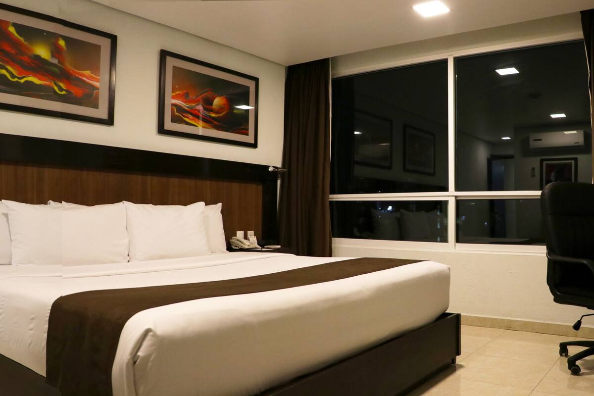 A hotel room with a large bed, a desk with a chair, a window with curtains, and two framed abstract paintings on the wall.