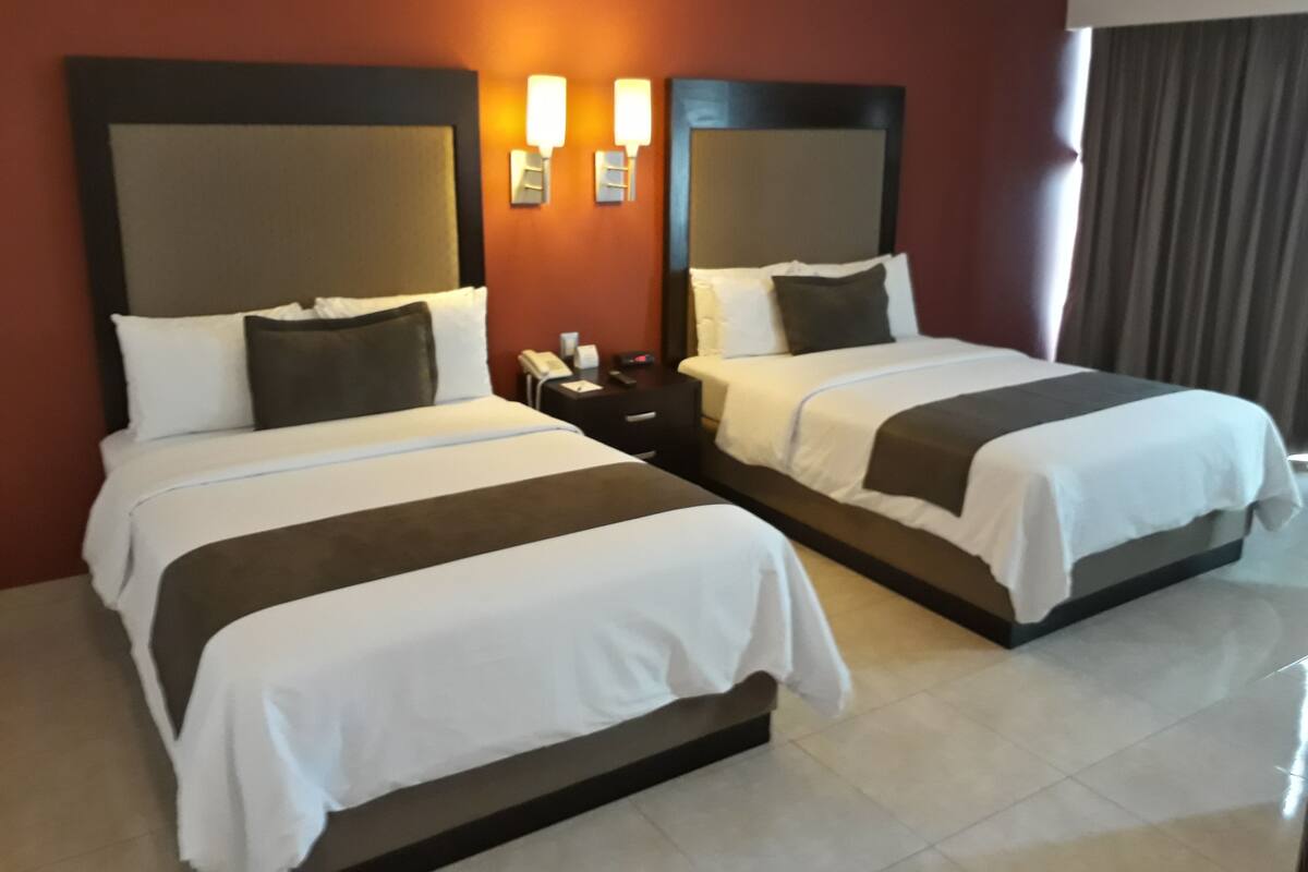A hotel room with two beds, each with a headboard, bedside tables, and wall-mounted lamps.