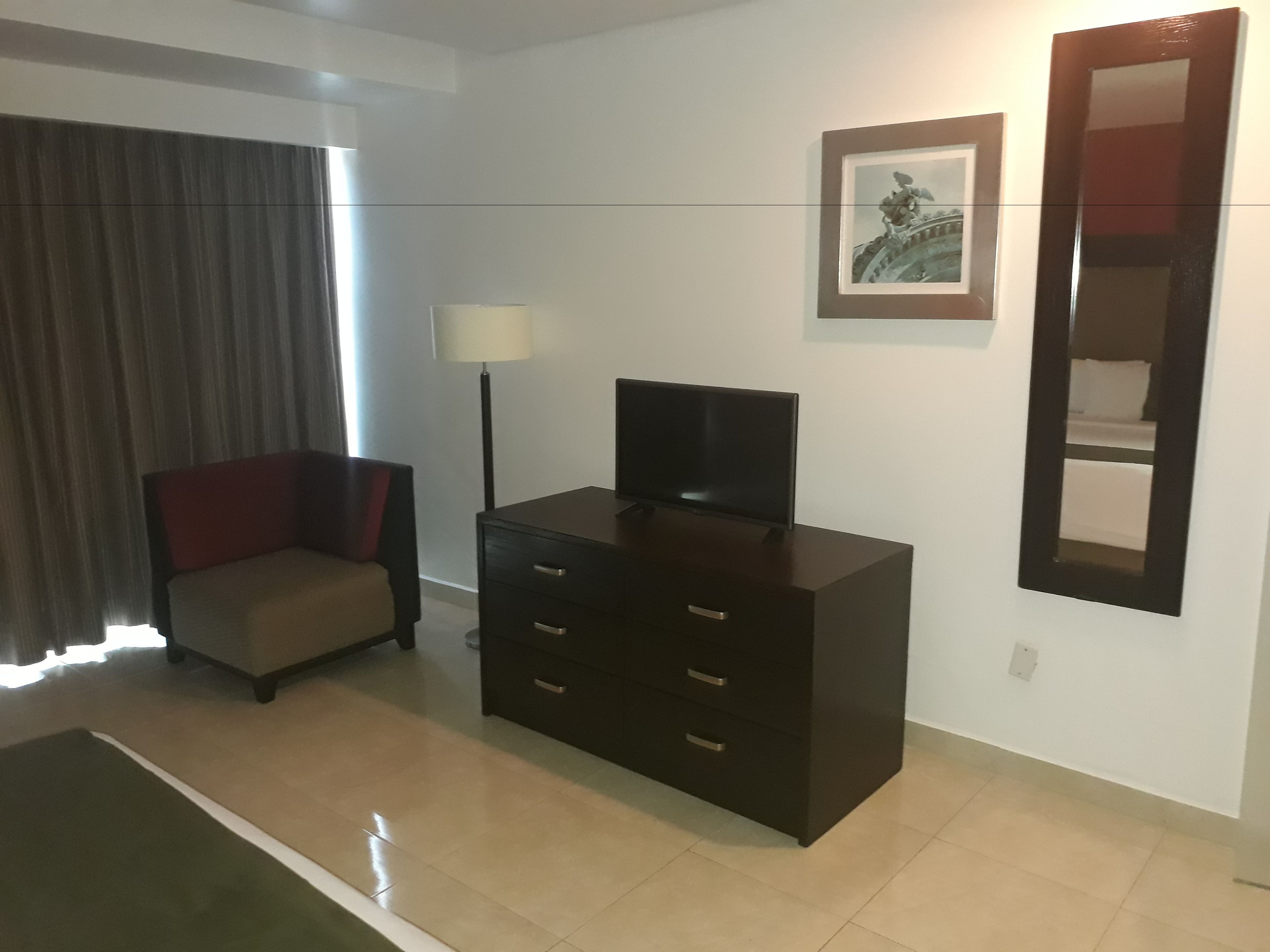 Une chambre d’hôtel avec une commode en bois foncé, un téléviseur posé dessus, une chaise, un lampadaire et un tableau encadré au mur.