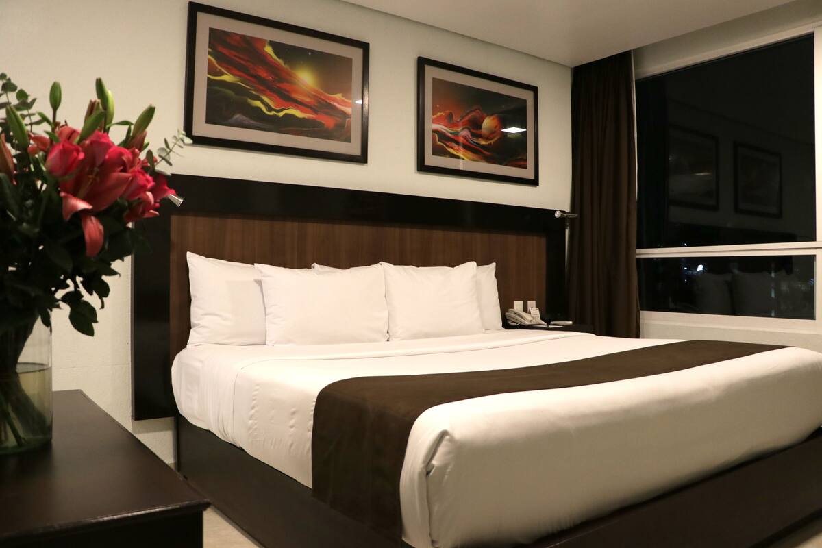 A hotel room with a large bed, a bedside table, a vase with flowers, and two framed artworks on the wall.