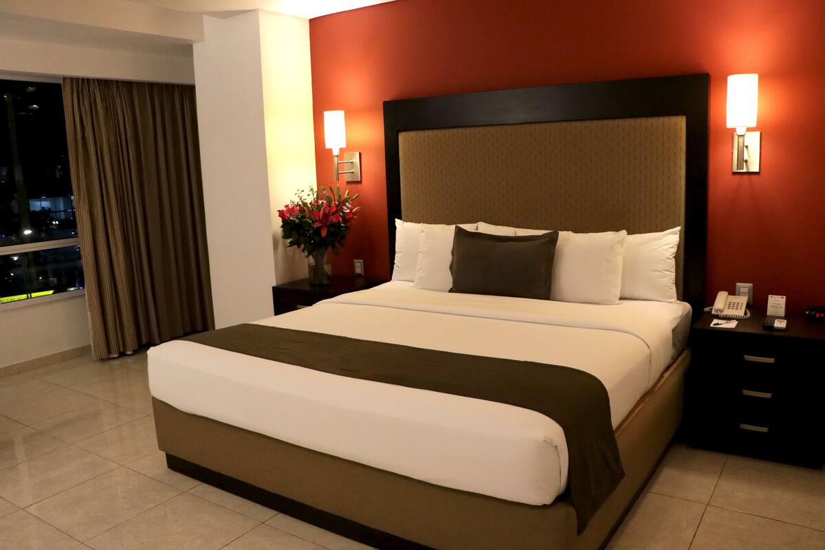 A hotel room with a large bed, a nightstand with a phone, and a view of the city at night.