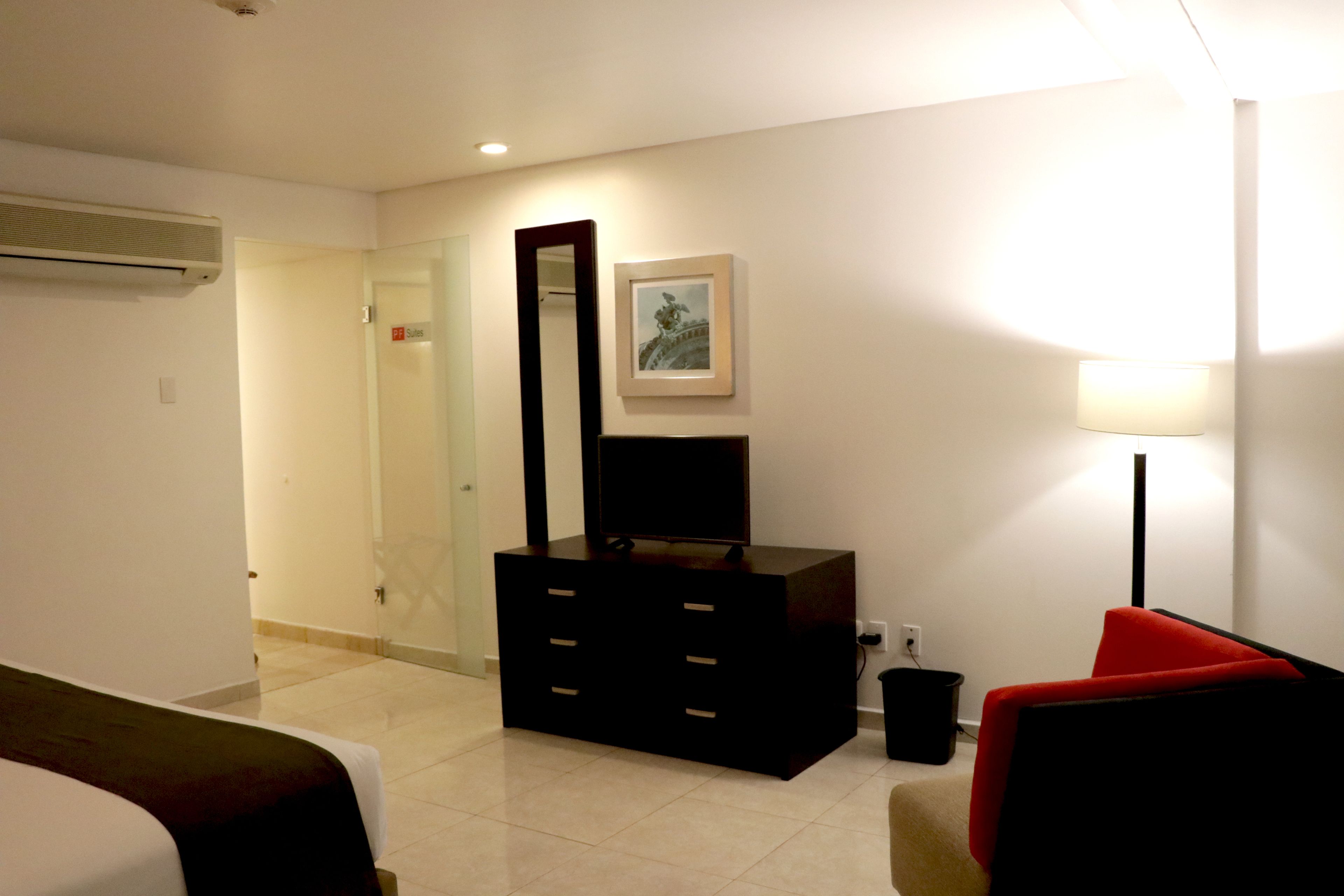 Une chambre d’hôtel avec un lit, une commode, un miroir, un tableau encadré, une chaise rouge et une lampe.